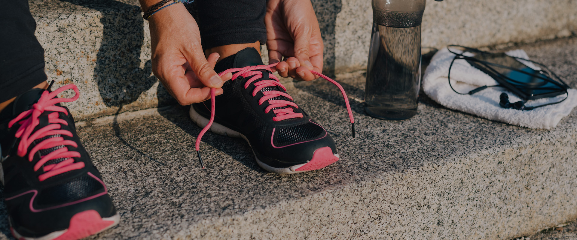 Woman tying pink laces of sport shoes closeup sitting on outdoors stairs with towel, bottle of water and phone with earphones before running training workout routine.