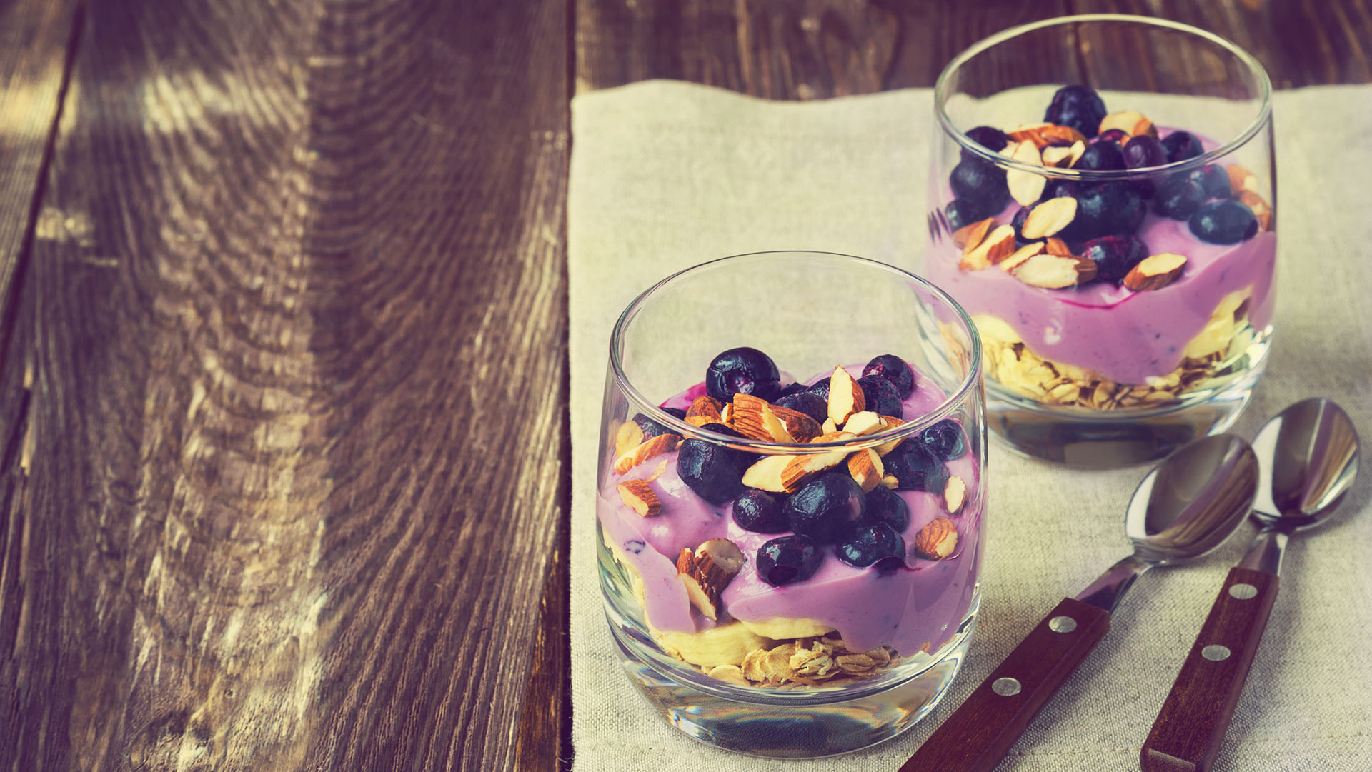Dessert of oatmeal, banana, yogurt, blueberries and almond in glass bowls on rustic wooden background. Vintage toned picture.