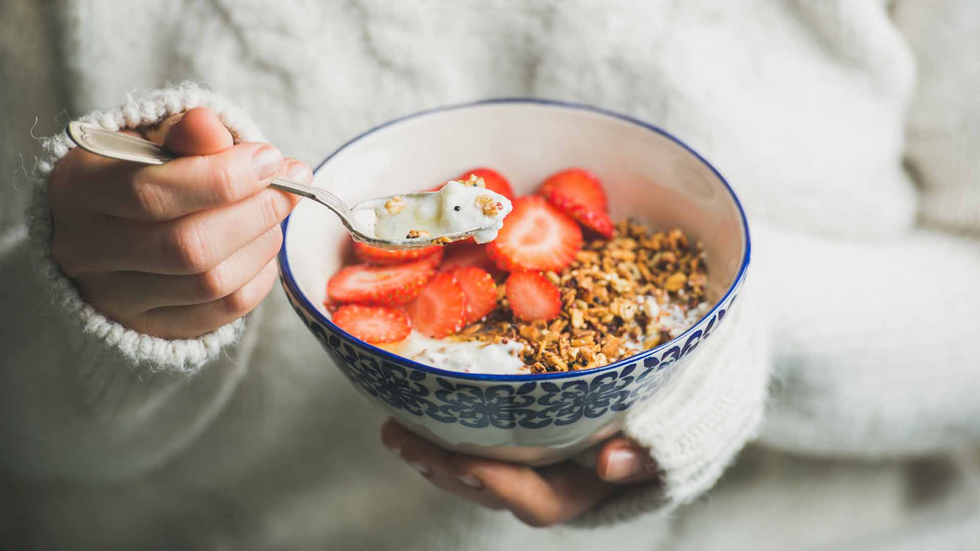 Healthy breakfast greek yogurt, granola and strawberry bowl in hands of woman wearing white loose knitted woolen sweater, selective focus. Clean eating, healthy, vegetarian, dieting food concept