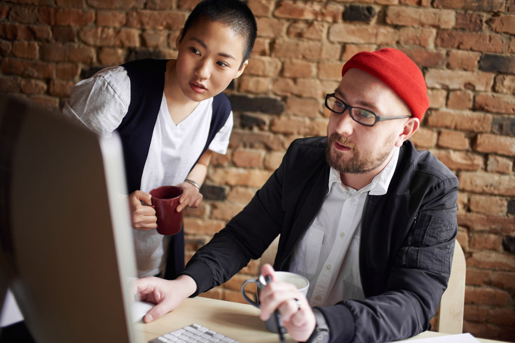 Male art director and his enthusiastic female coworker looking at computer screen attentively against brick wall background