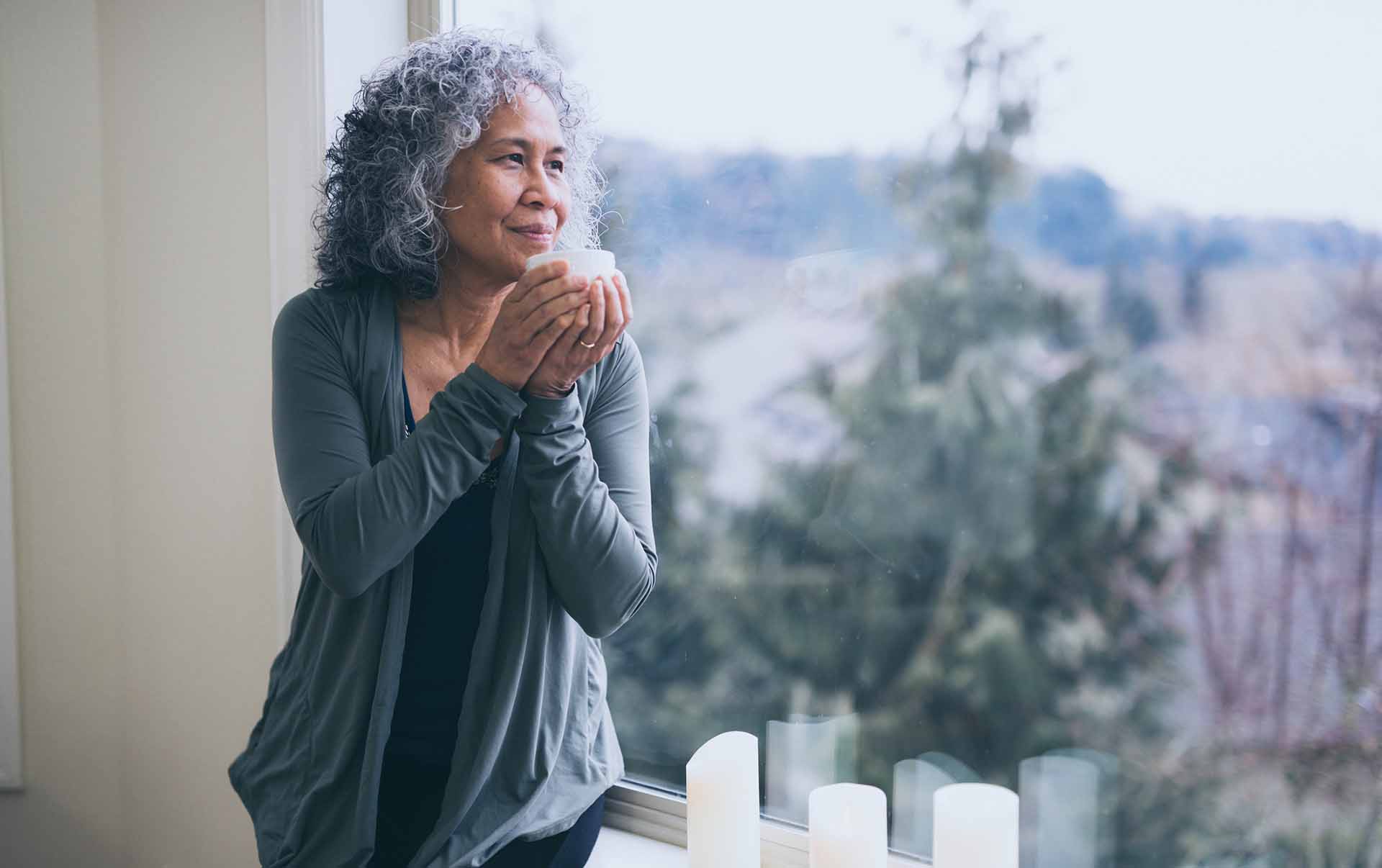 A beautiful ethnic older woman sits in her window sill and looks out contemplatively while she holds a cup of tea.