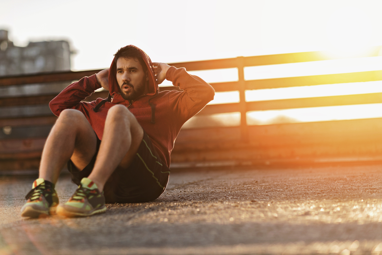 Picture of a young athletic man doing sit ups sitting on a the rough ground. He is wearing a red sweater with the hood up. In the background is a brown fence and the sun is setting.