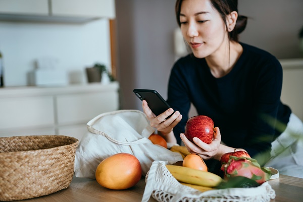 Beautiful smiling young Asian woman grocery shopping online with mobile app device on smartphone, checking her fresh fruits delivery. With a reusable mesh bag full of fresh and healthy organic fruits on table. Responsible shopping and technology concept