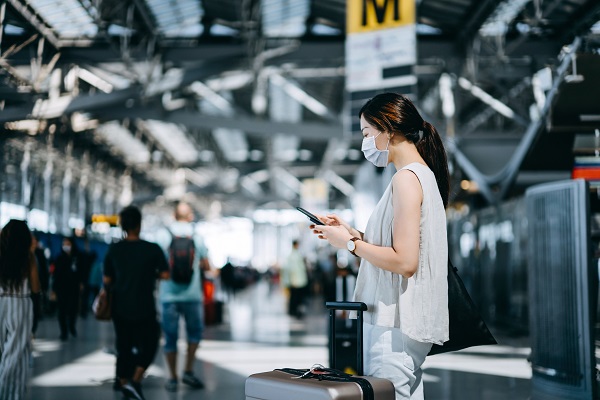 Essential travels during Covid-19 pandemic. Young Asian woman with protective face mask checking in online with her smartphone in airport terminal, with suitcase by her side