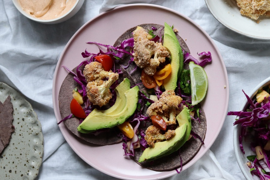 A plate of cauliflower tacos with creamy avocado slices and crisp purple cabbage on blue corn tortillas, accented with halved cherry tomatoes and a lime wedge, alongside a bowl of chipotle cream.