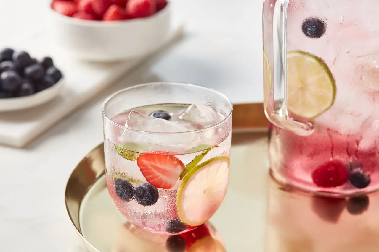 A refreshing mocktail with ice, sliced strawberries, blueberries, and lime in a glass, set on a golden tray beside bowls of fresh berries, with a pitcher of the same drink in the background.