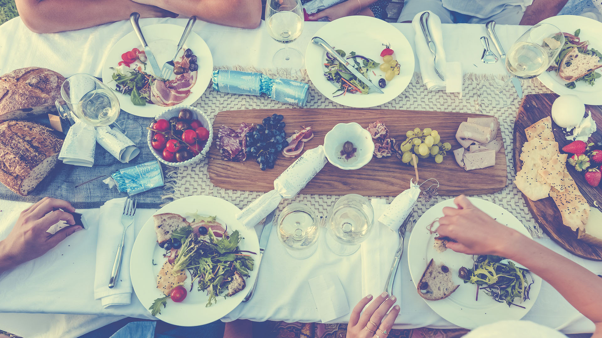 Group of people with broken Christmas crackers. There is food on the table outdoors in a December Summer. There is Cheese meat and wine. Could be a family. High angle view. You can just see a Santa hat