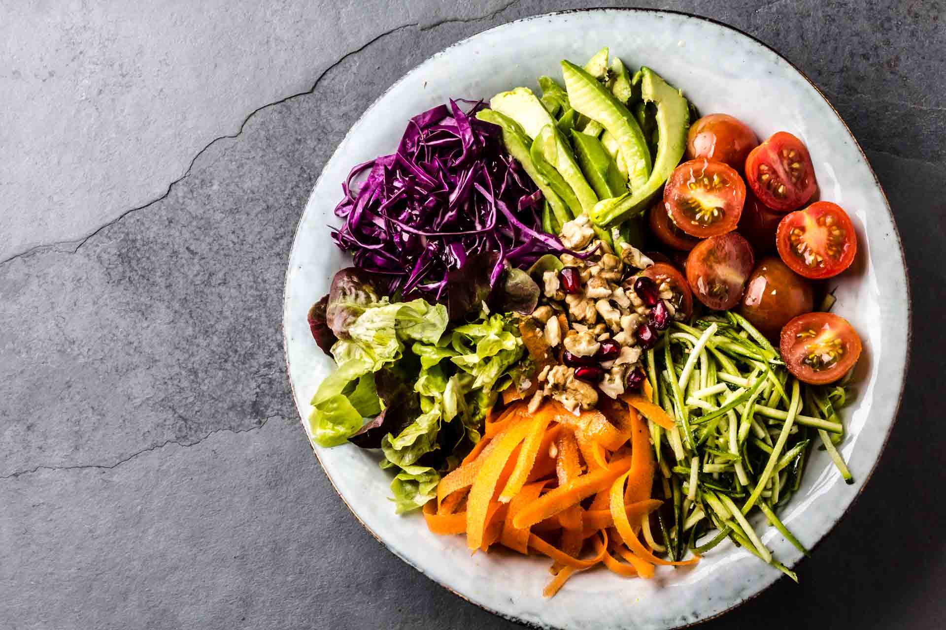 Vegan buddha bowl. Bowl with fresh raw vegetables - cabbage, carrot, zucchini, lettuce, watercress salad, tomatoes cherry and avocado, nuts and pomegranate on slate background
