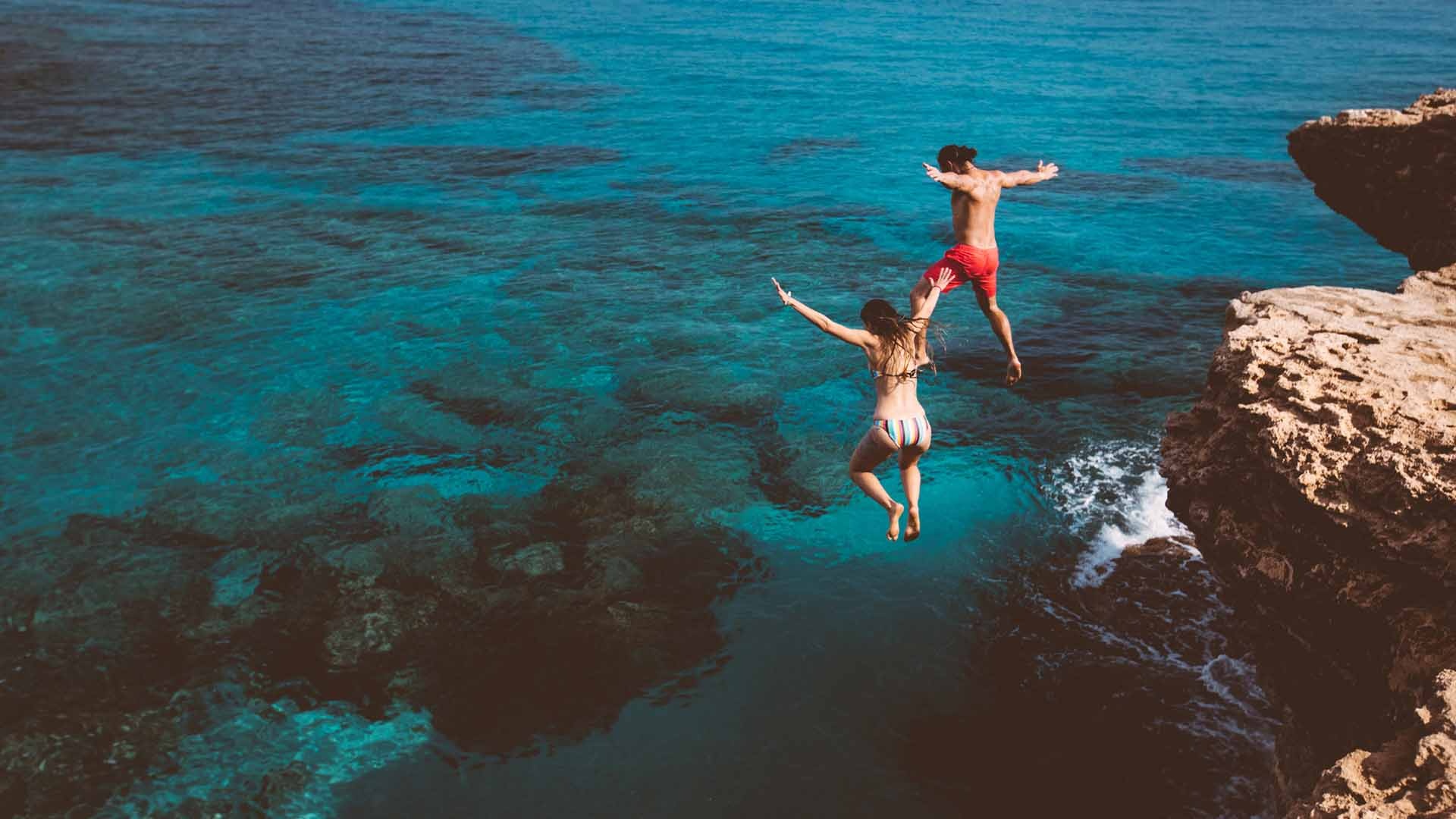 Young active man and woman diving from high cliff into tropical island blue sea water