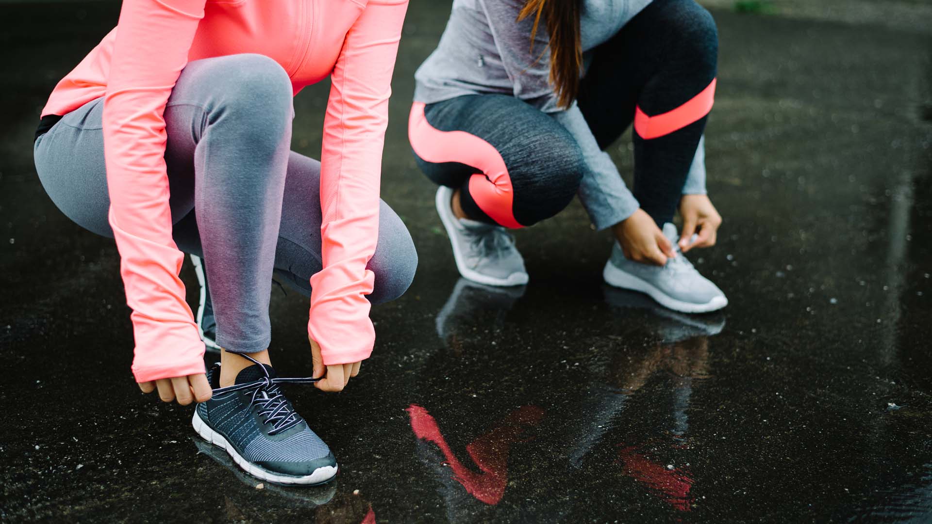 Urban athletes lacing sport footwear for running over asphalt under the rain. Two women getting ready for outdoor training and fitness exercising on cold winter weather.