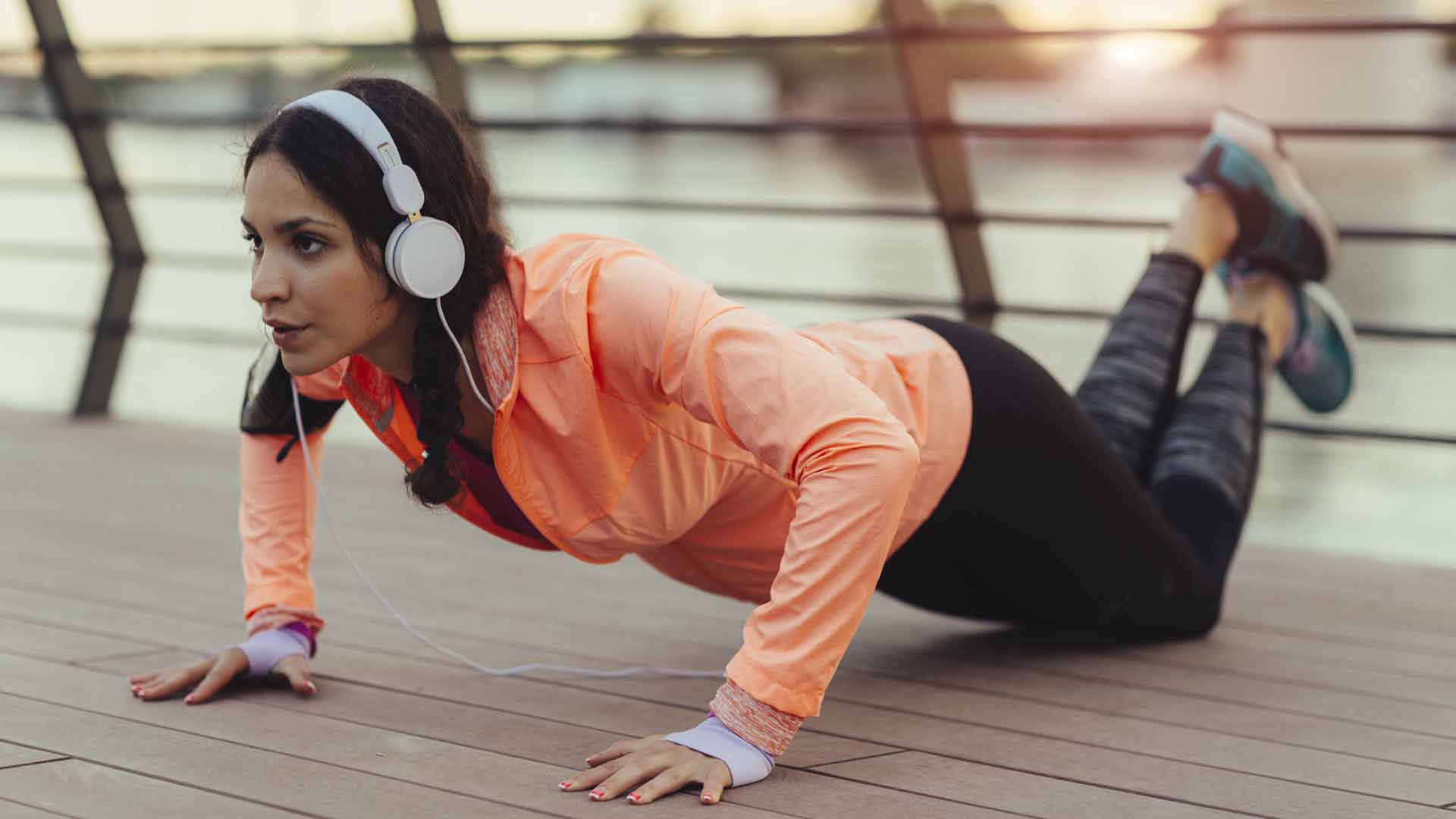 Young woman exercising near river at sunset. She is wearing modern sporty clothing. Listening music with big white headphones and doing pushups.