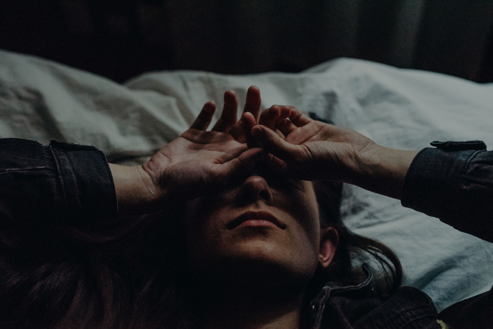 Young adult woman lying on a bed with crinkled white sheets in a dark cabin with little window light with her hands and fingers relaxed on her face hiding her eyes from the camera