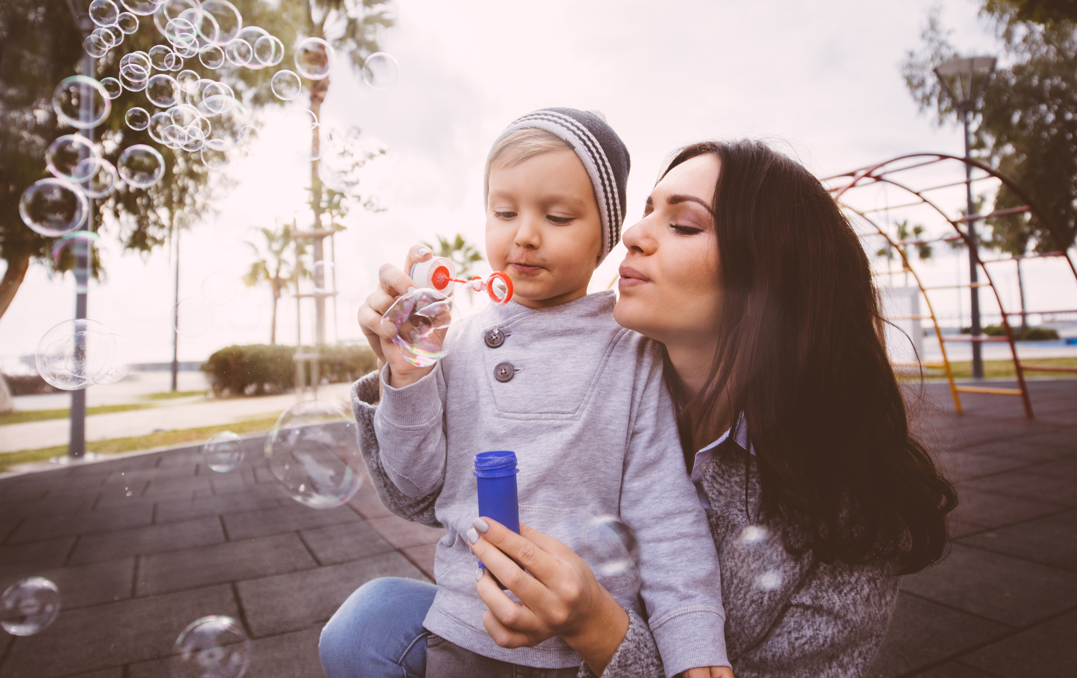 Shot of young blond boy blowing bubbles with the help of his mum at the playground