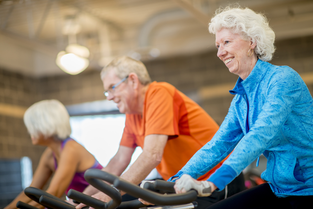 A group of senior adults are working out together at a spin class. They are sitting on their stationary bikes and and cycling together to the music.