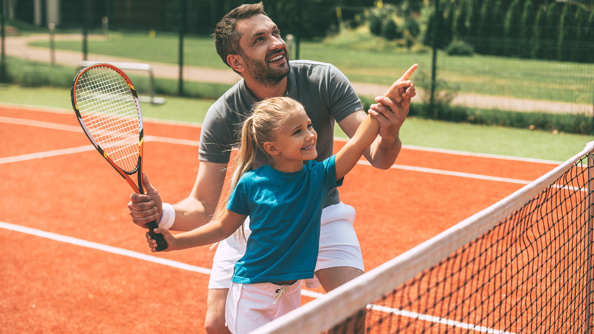 Cheerful father in sports clothing teaching his daughter to play tennis while both standing on tennis court