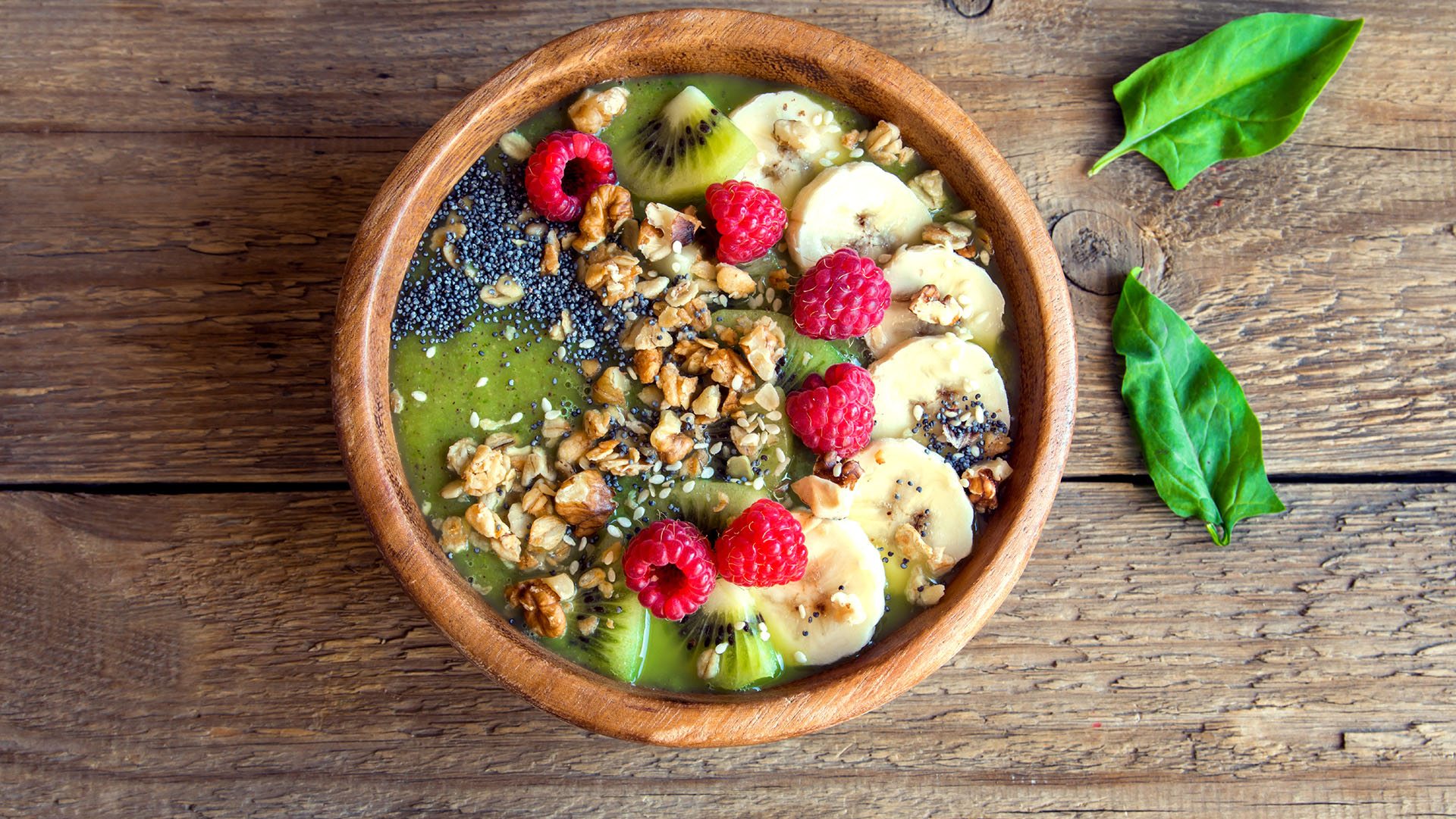 Healthy breakfast green smoothie bowl topped with fruits, nuts, berries and seeds over rustic wooden background