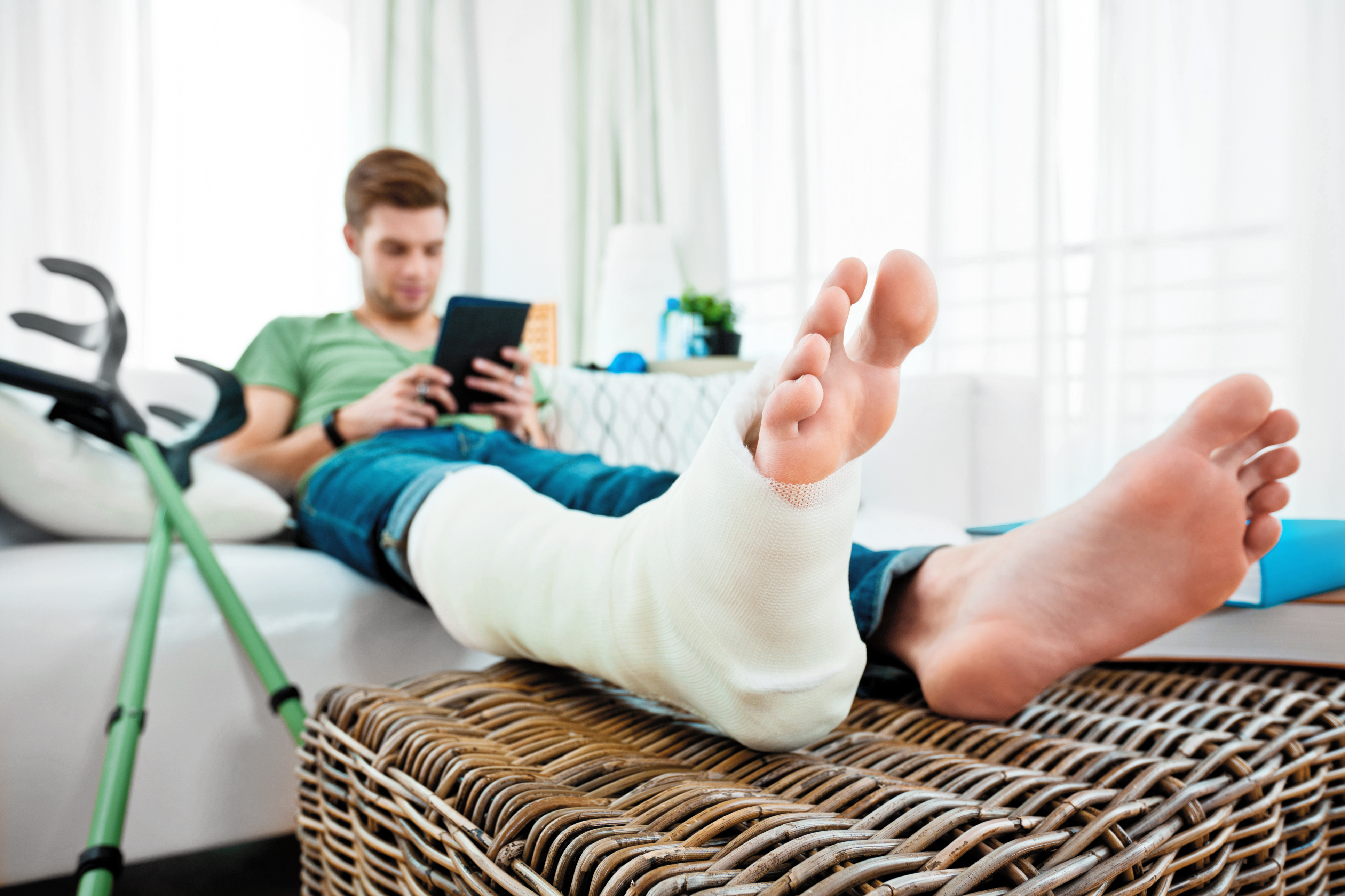 Young man with broken leg sitting on sofa at home and reading e-book. Focus on foreground.