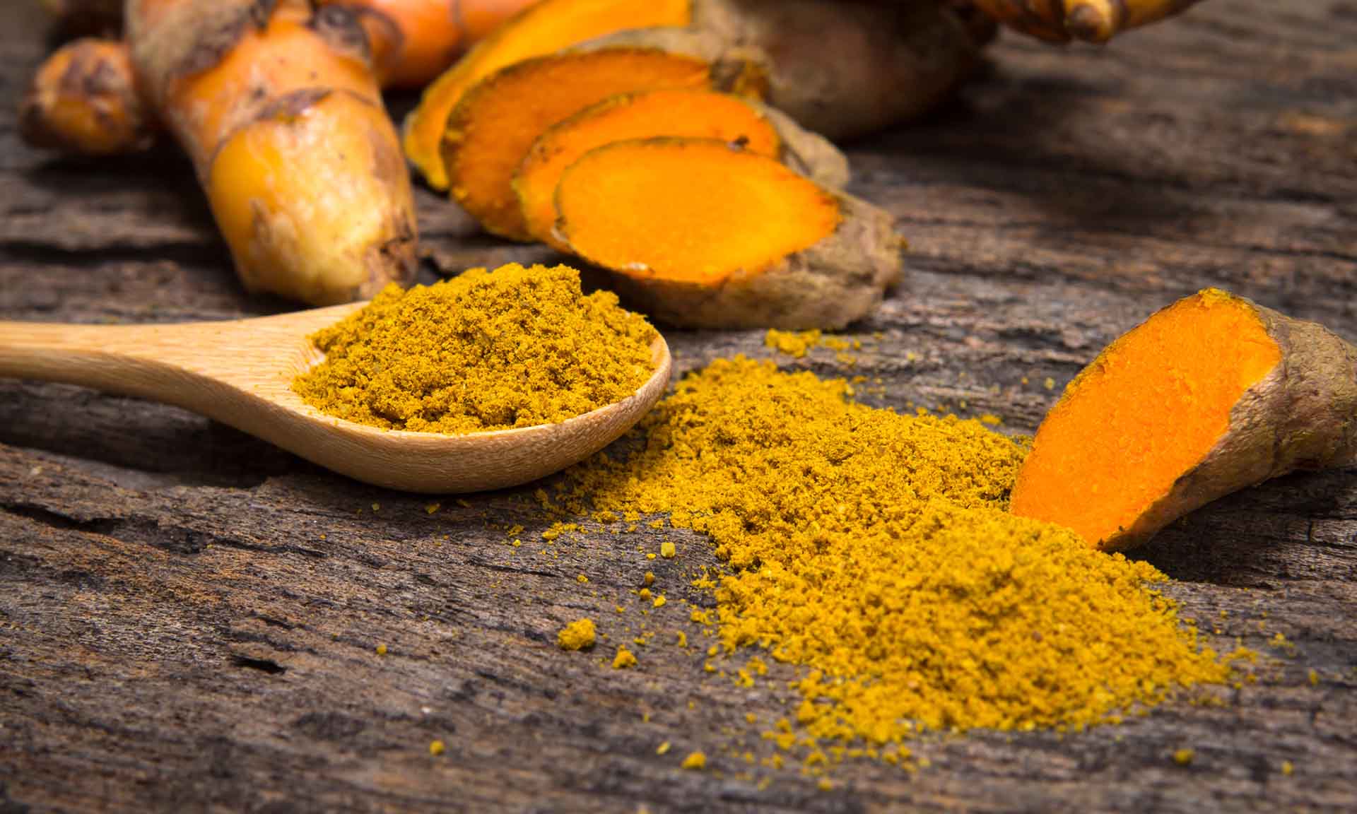 the turmeric powder in spoon and roots on wooden plate