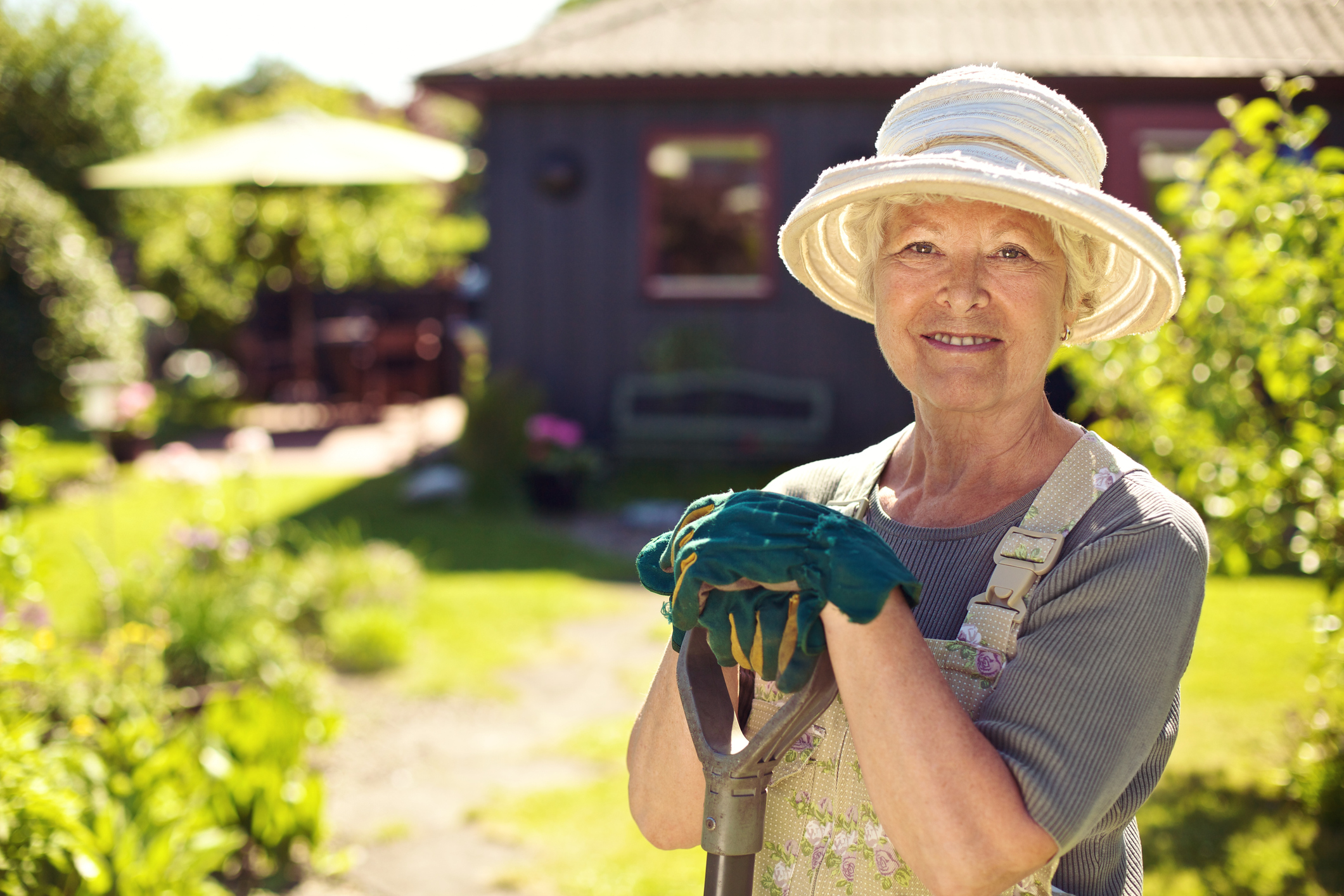Portrait of senior woman wearing hat with gardening tools outdoors. Elder woman standing with shovel in her backyard garden