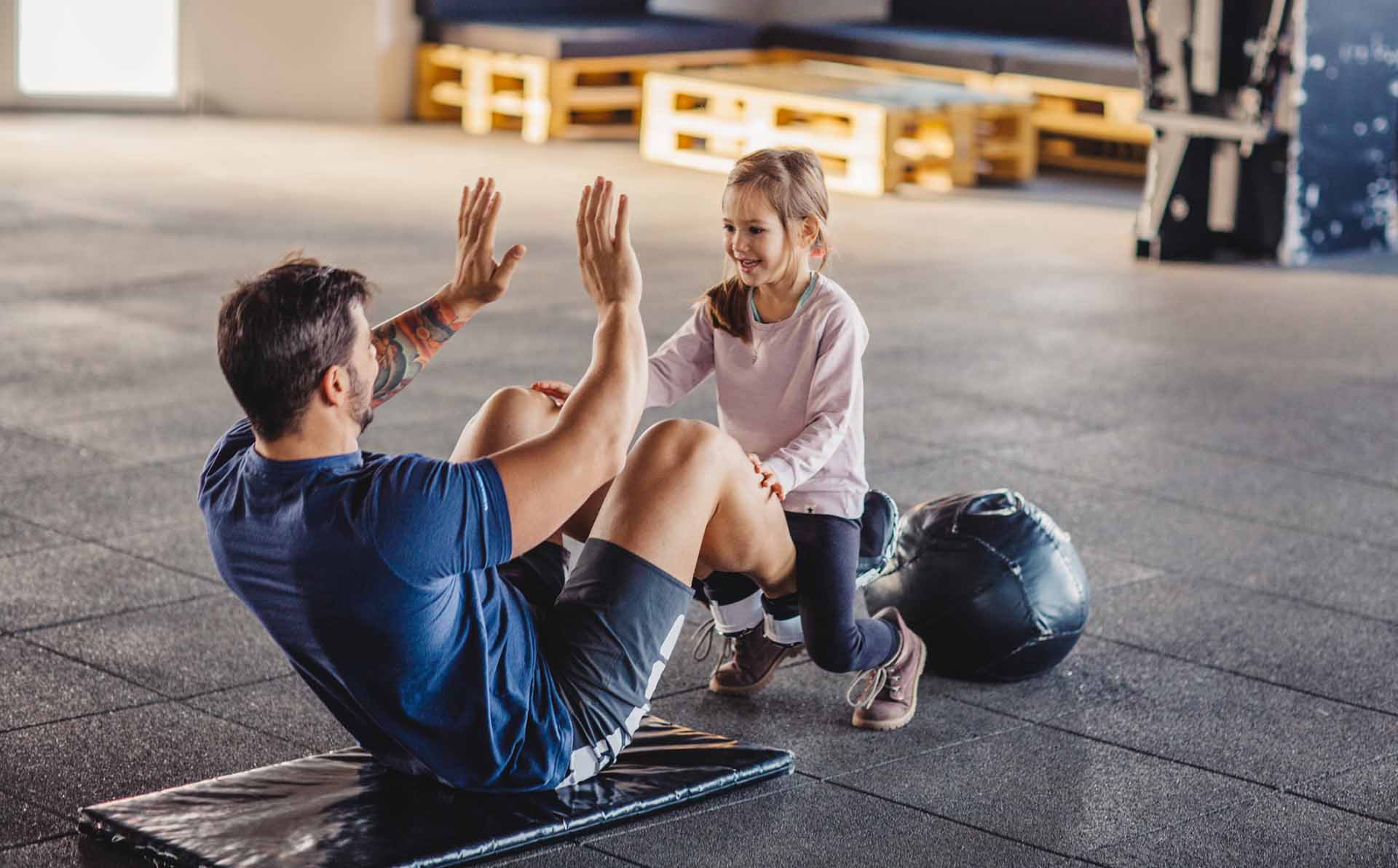 Father doing sit-ups with daughter sitting on his legs . They are in gym, exercising together.