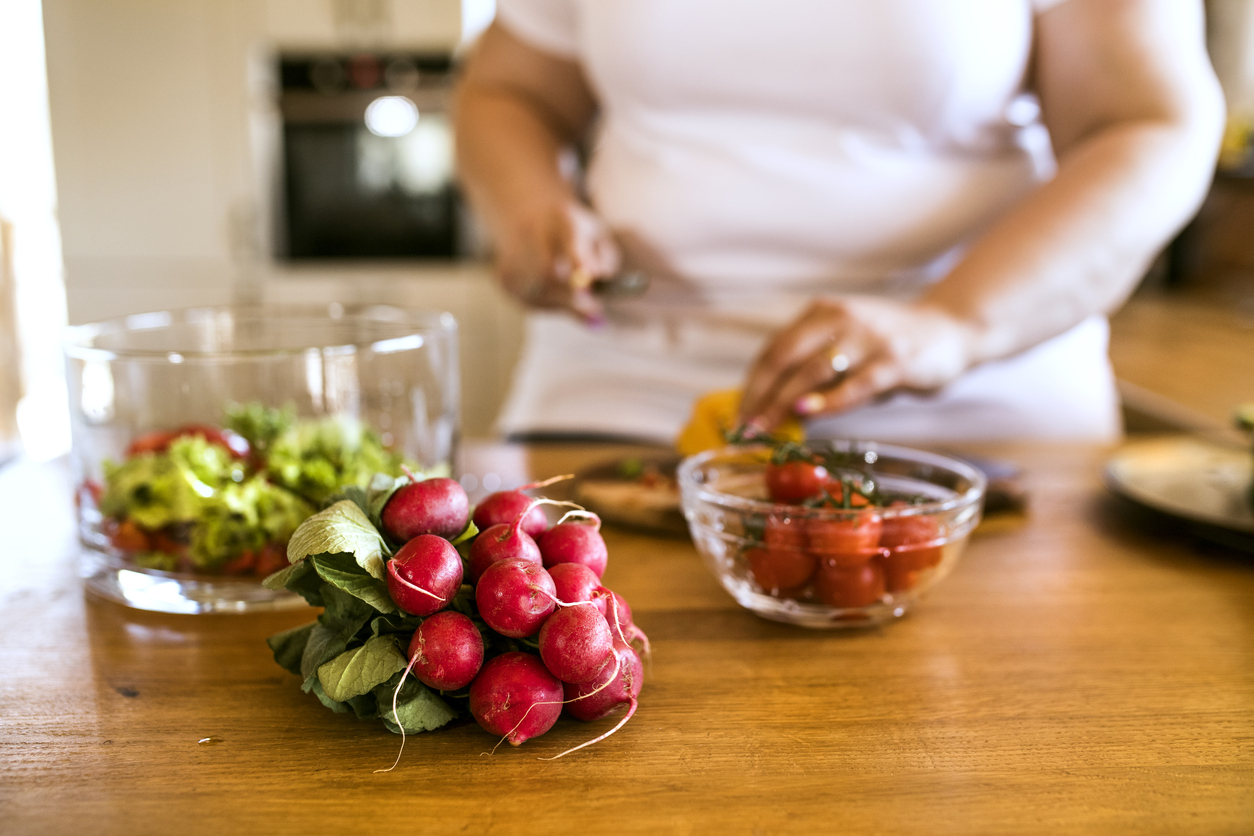 Unrecognizable overweight woman in white t-shirt at home preparing a delicious healthy vegetable salad in her kitchen.