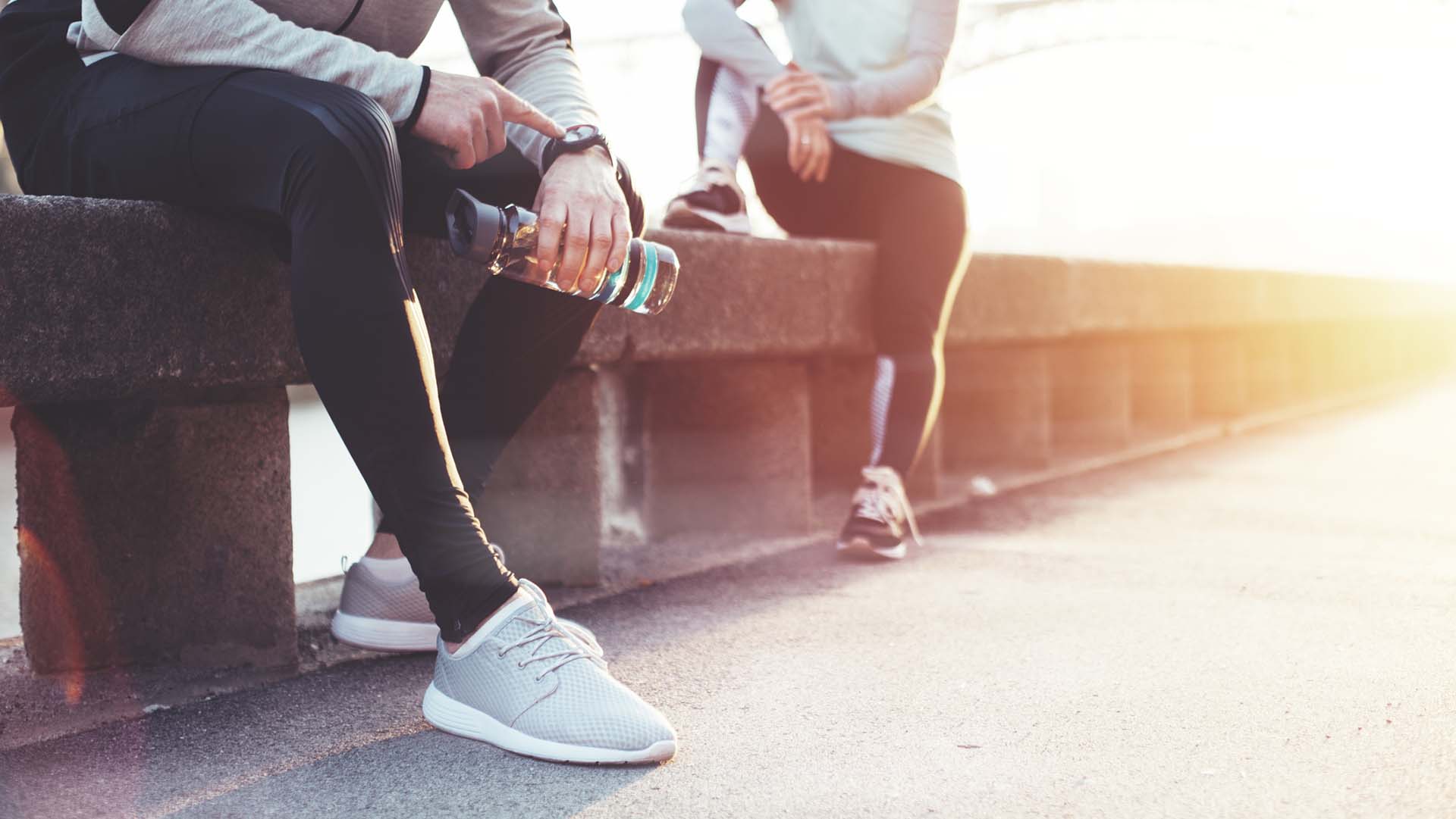 Couple of athletes resting after workout session on the street. Tired man with bottle tracking time with his watch and woman relaxing on the bench