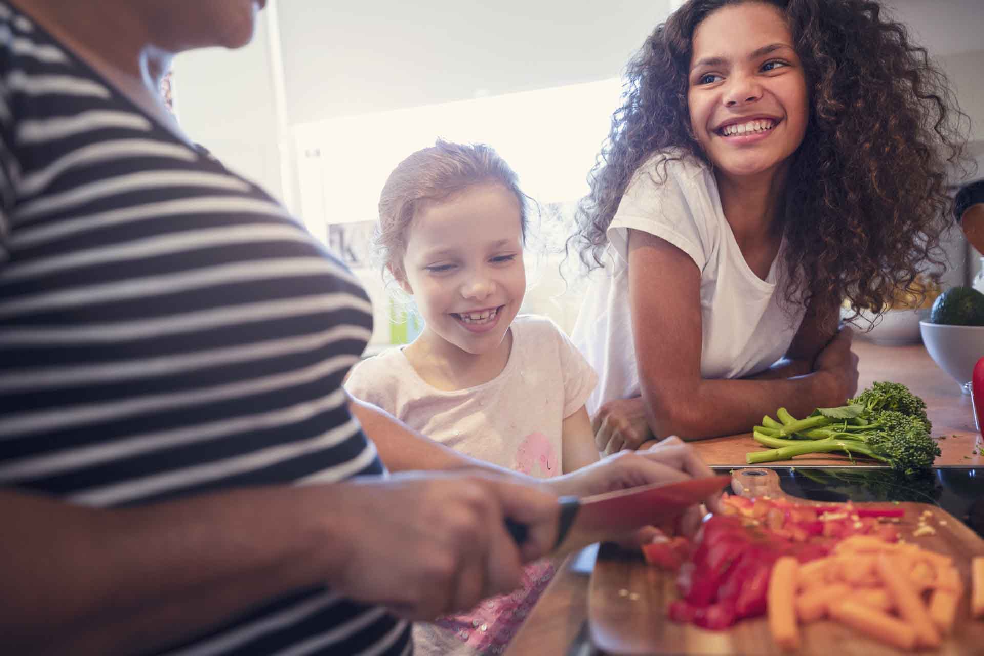 Mother and children cooking in the kitchen. They are cooking a healthy meal and talking. The mother may be teaching her daughters. There are vegetables on a cutting board in the foreground including peppers, carrots and tomatoes with an empty pan.