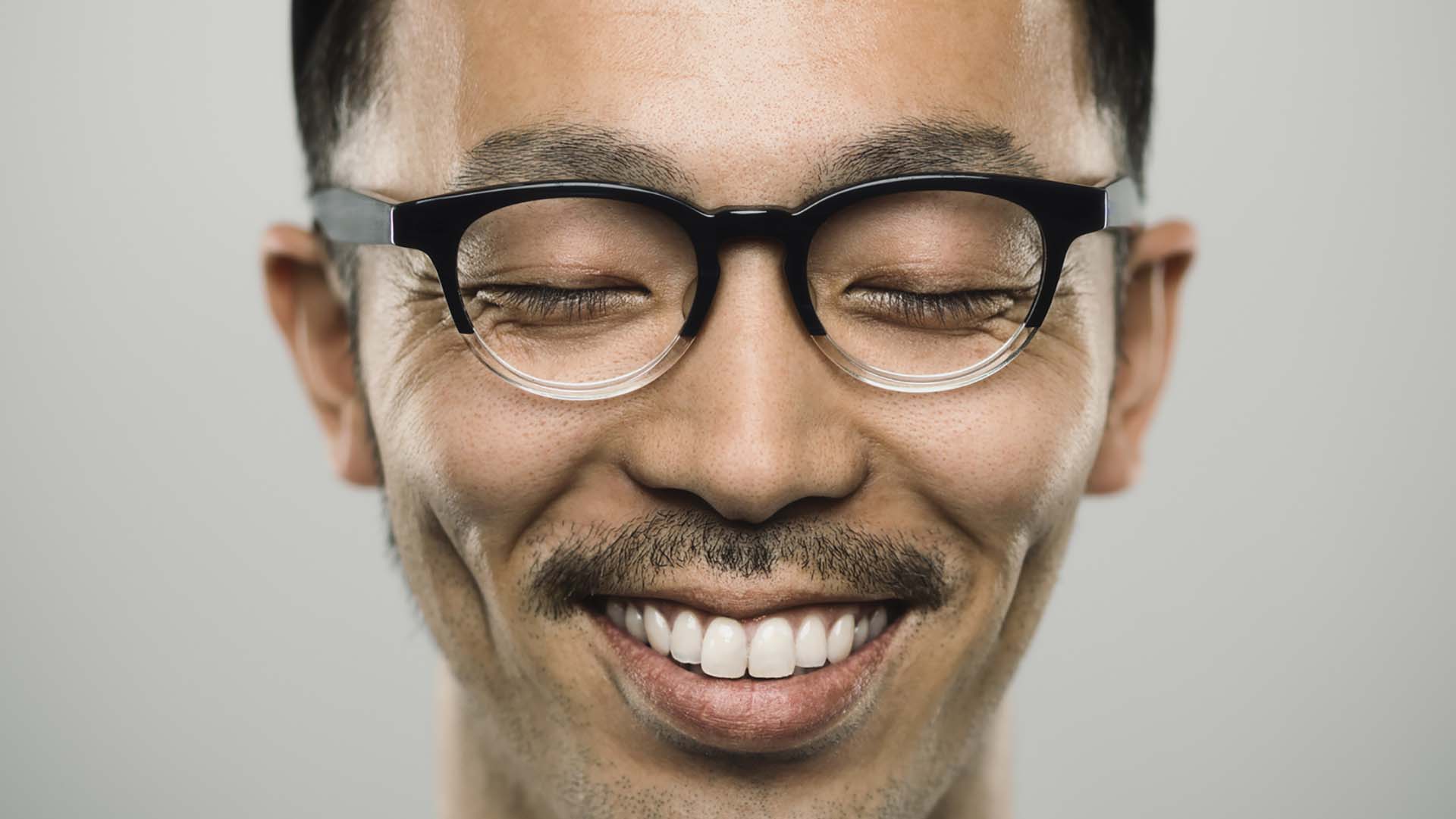 Studio portrait of a japanese young man with closed eyes and very big smile. The man is around 30 years and has short hair and casual clothes, wearing a baseball cap and glasses. Vertical color image from a medium format digital camera. Sharp focus on eyes.