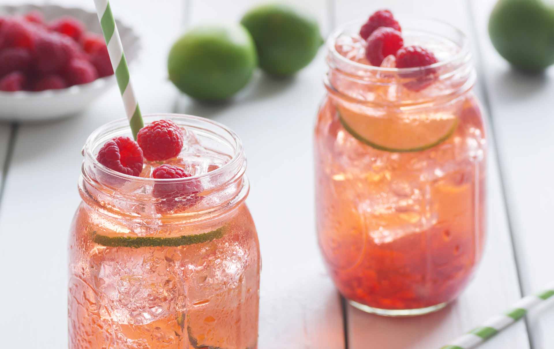 Raspberry lime iced tea in glasses with straws. Ingredients for making tea - including fresh limes and berries in a bowl - are on the white picnic table in the background.