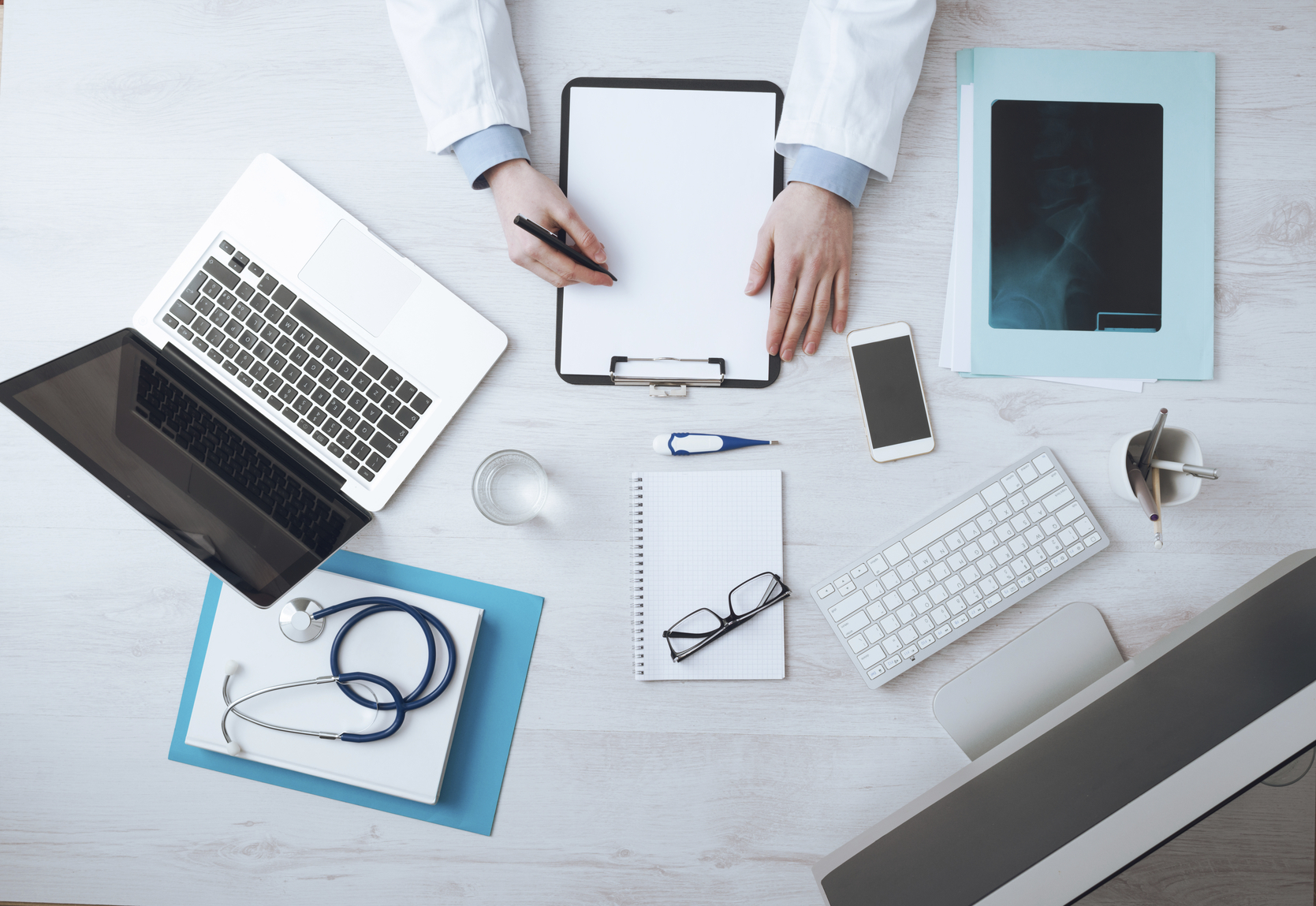 Professional doctor writing medical records on a clipboard with computer and medical equipment all around, desktop top view