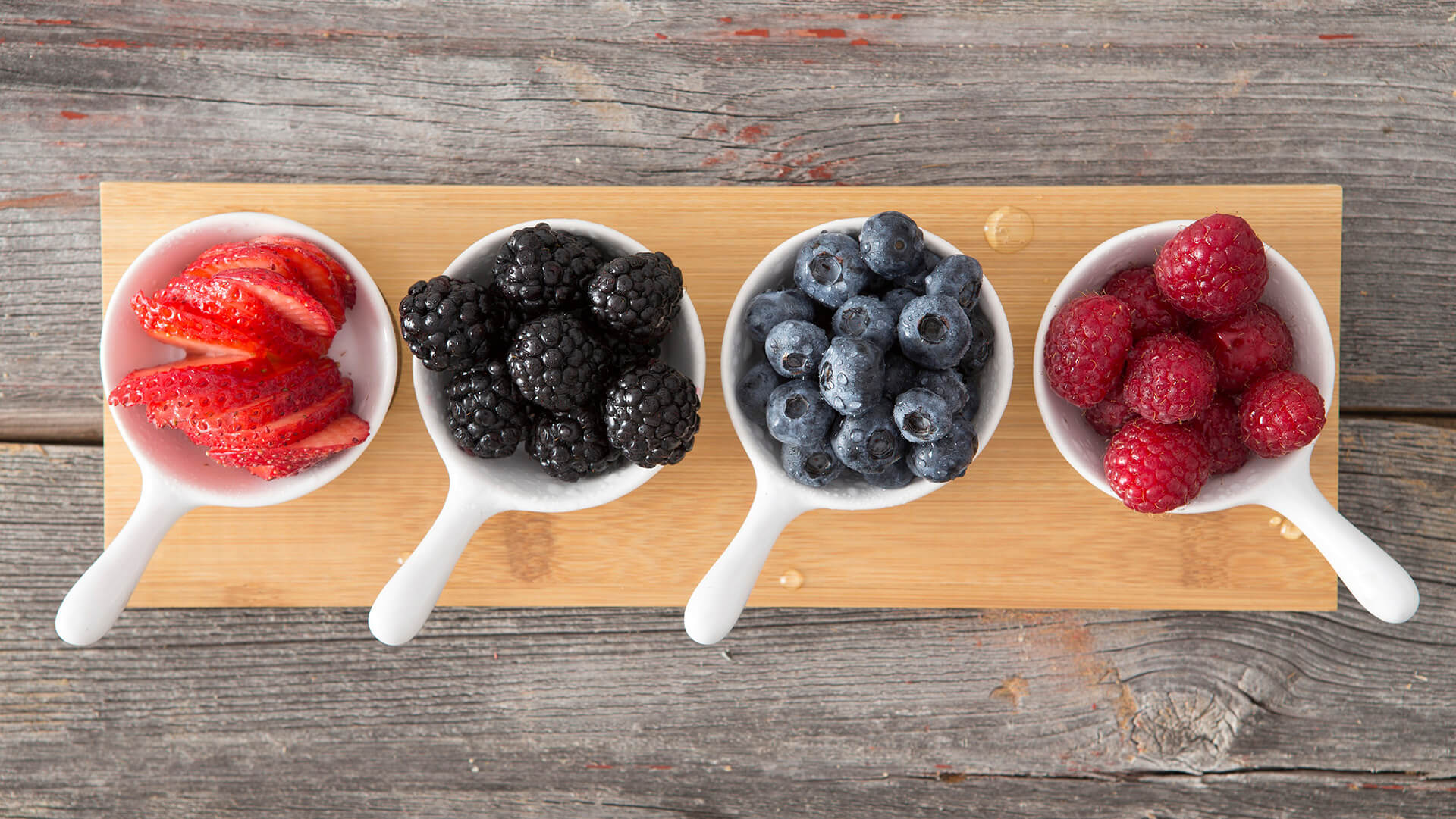 Taster dishes of assorted autumn berries viewed from above arranged on a wooden board on a rustic, kitchen table including whole fresh blueberries, blackberries, raspberries and sliced strawberries
