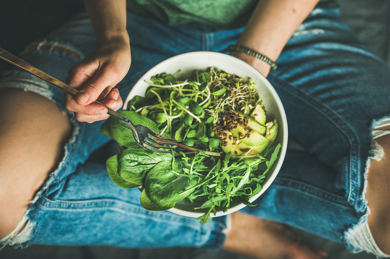 Green vegan breakfast meal in bowl with spinach, arugula, avocado, seeds and sprouts. Girl in jeans holding fork with knees and hands visible, top view. Clean eating, dieting, dieting food concept