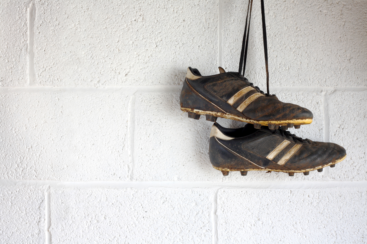 A pair of muddy black football boots hang up in a white-walled changing room. There is room for copyspace.