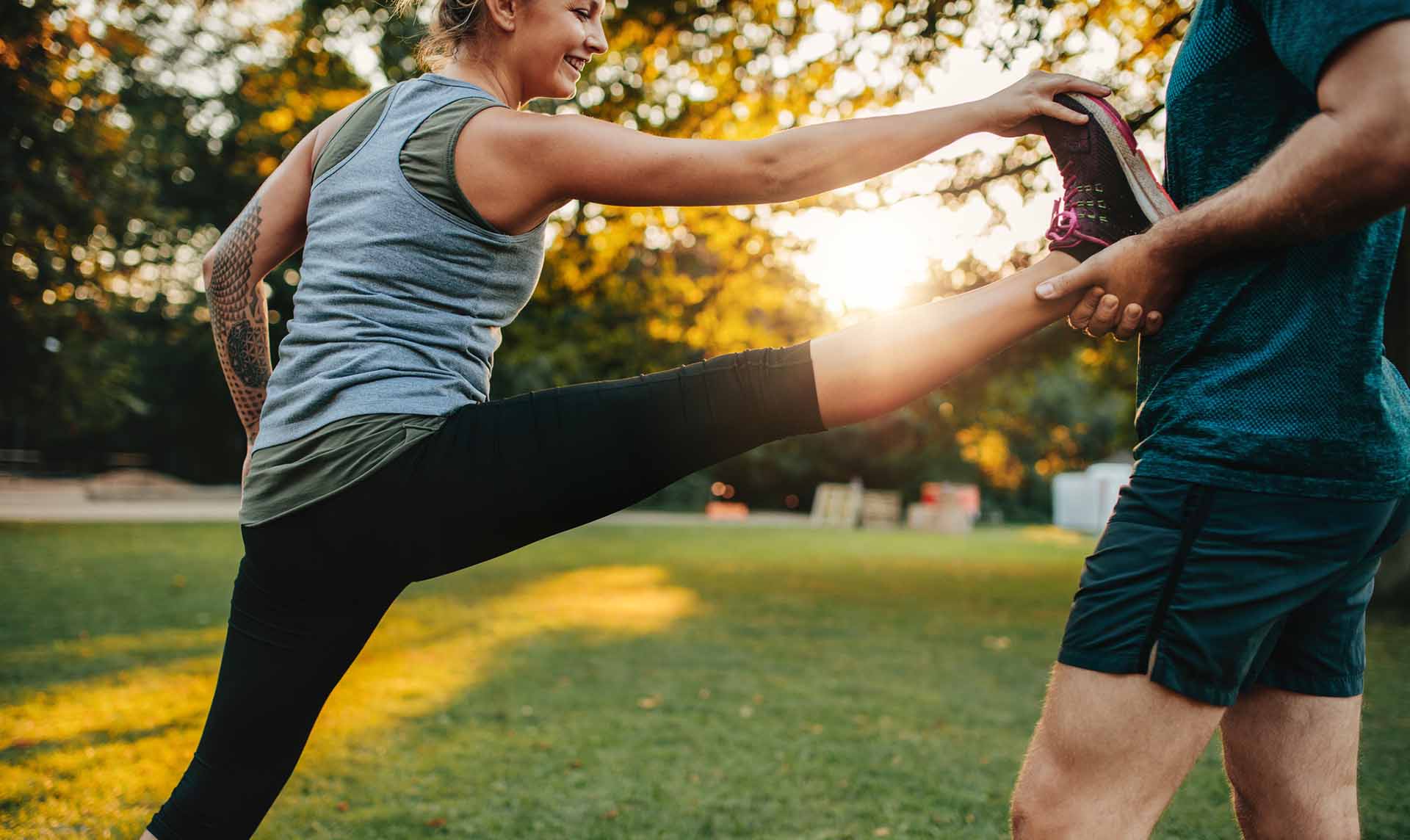 Fit young woman doing stretching exercises with the help of a personal trainer in park. Coach holding leg of female.