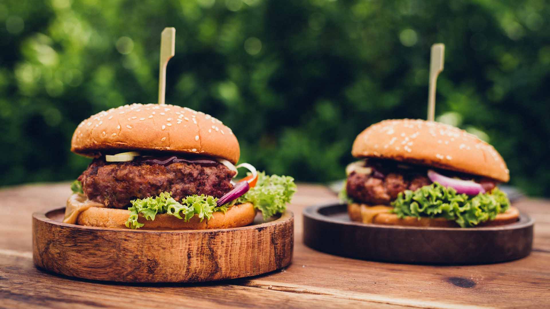 Low angle shot of two delicious gourmet burgers with high quality beef and fresh ingredients presented on wooden platters outdoors in a lush back yard