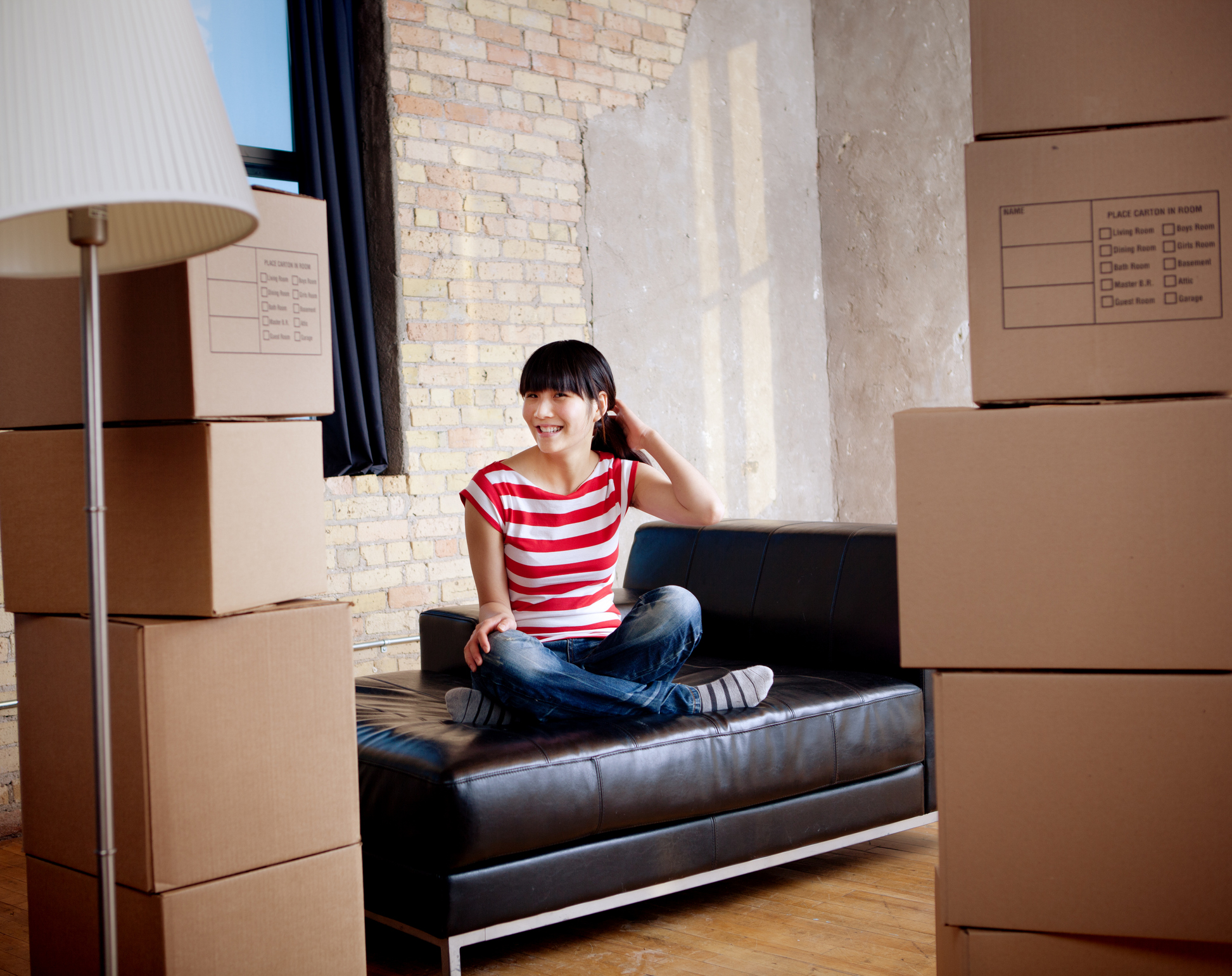 Subject: Horizontal view of a young Asian woman sitting cross legged on a sofa, surrounded by moving boxes. The interior suggests that she is happy that she has just moved into an urban loft apartment or condo.