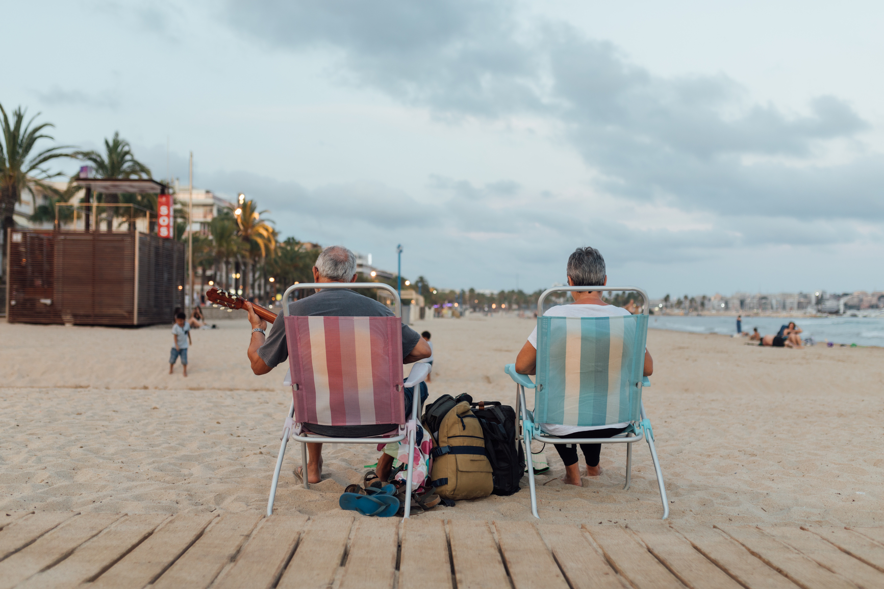 Retired couple playing guitar on a summer afternoon at the beach. They wear comfortable and simple clothes, they are barefoot. They are sitting on beach chairs at the edge of the road to the sea. We see them from behind.