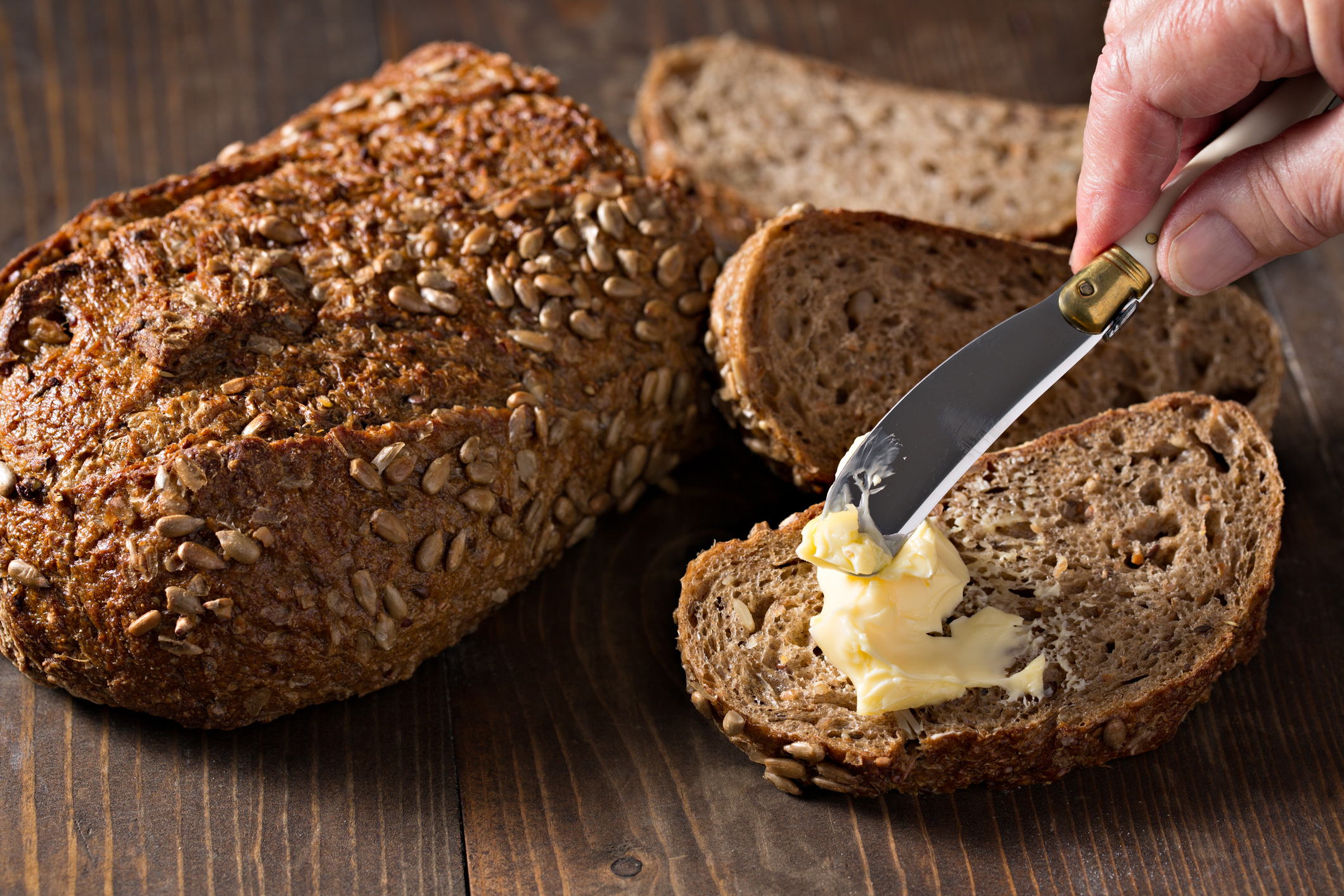 A high angle, close up photograph of a hand spreading some fresh butter on a slice of healthy whole grain bread.