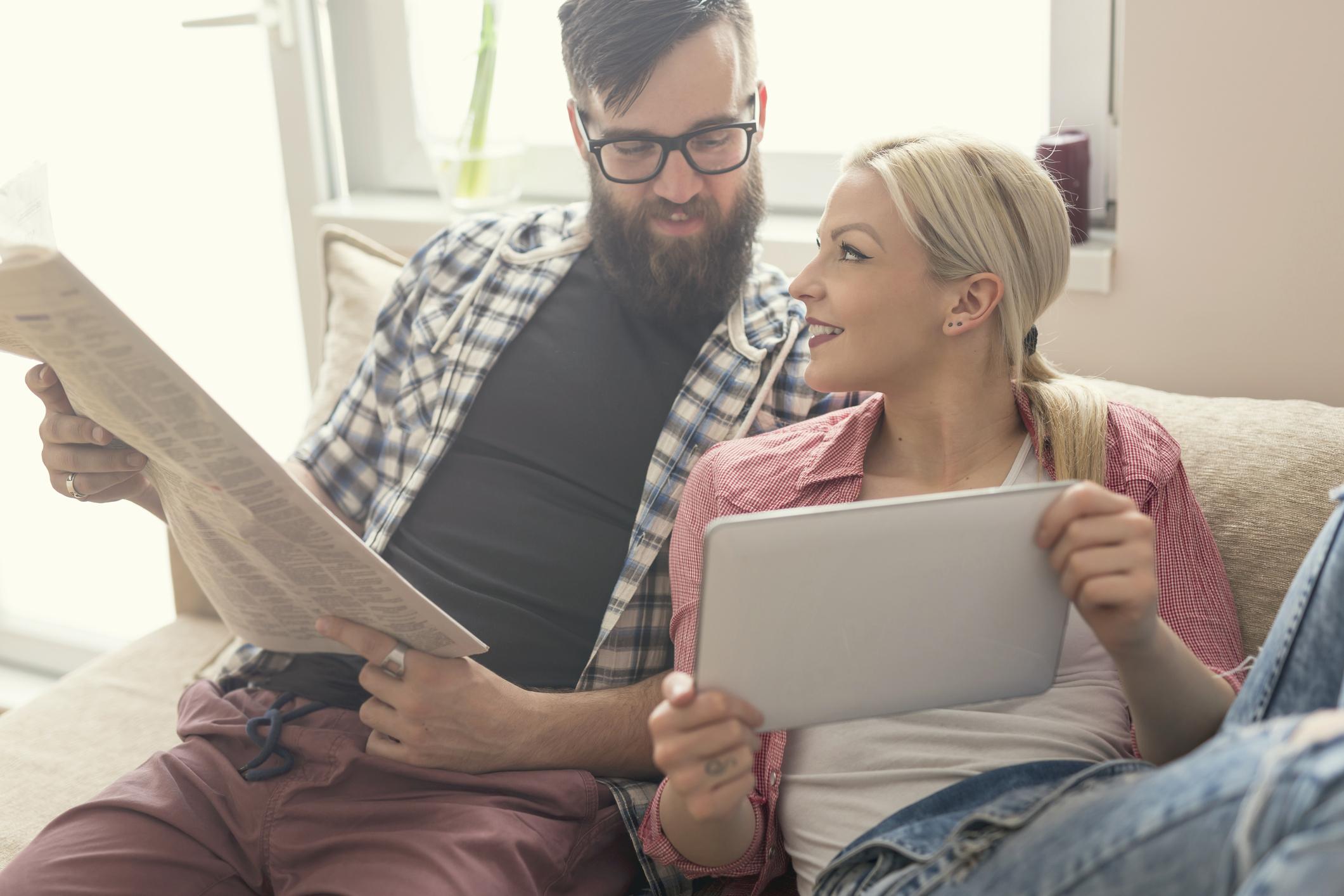 Young couple in love sitting on a couch in their apartment, enjoying their free time, reading newspaper and surfing the web on a tablet computer