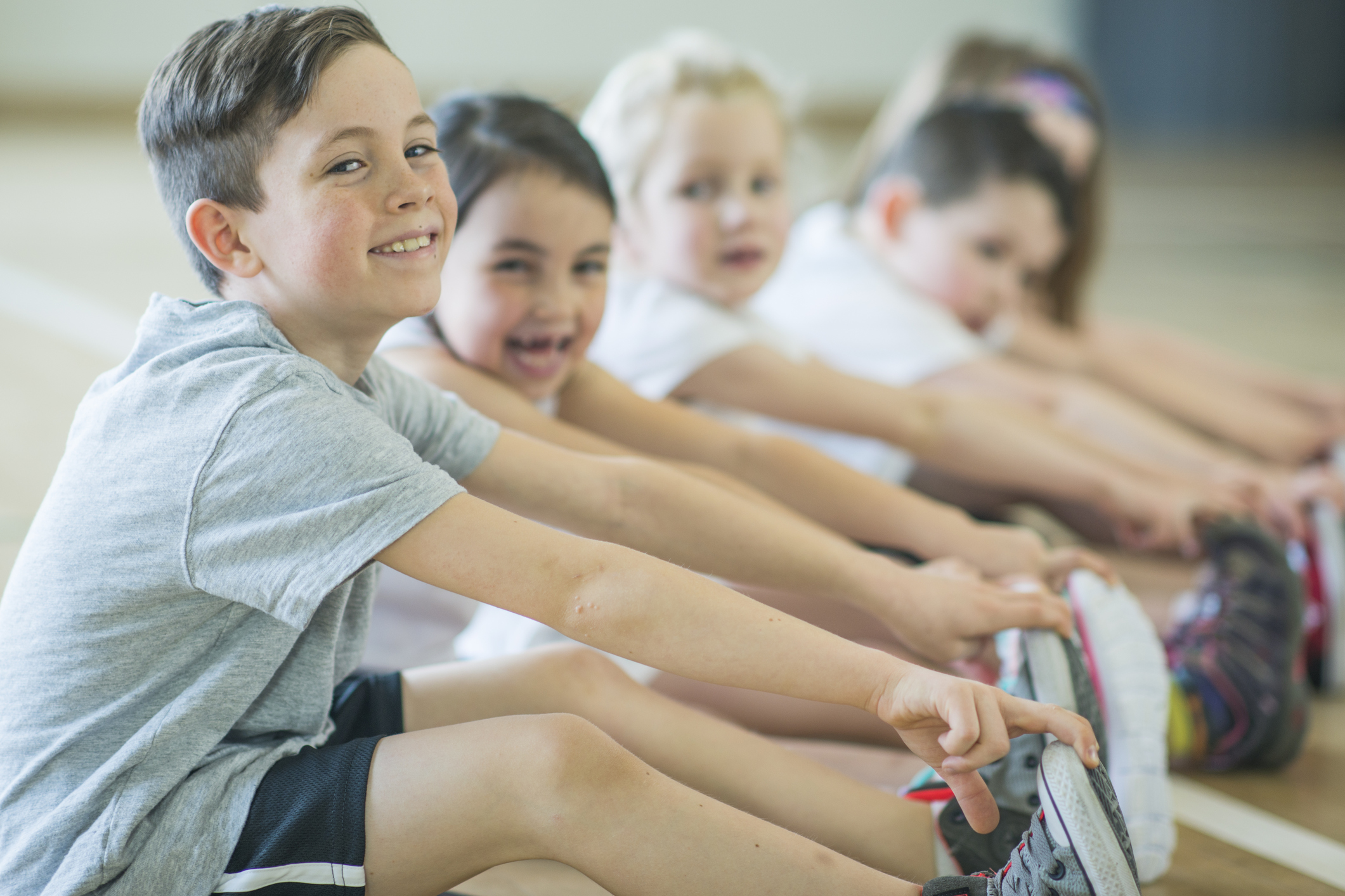 A group of Caucasian elementary school students are indoors in a gym. They are wearing casual sports clothing. They are sitting on the floor and touching their toes. They are doing stretching exercises.