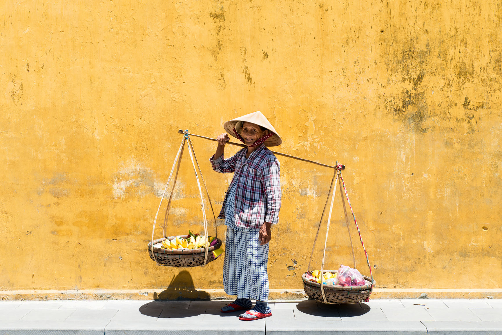 Side view of old woman in traditional Vietnamese hat carrying baskets full of bananas along sidewalk against grunge dirty yellow wall.