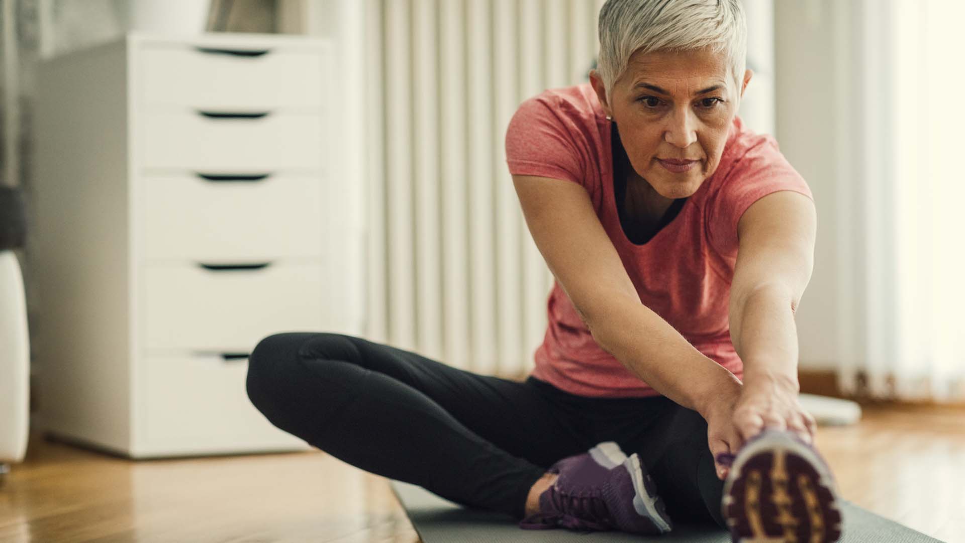 Mature woman exercising at home. Sitting on exercise mat and working stretching exercise , stretching her legs.
