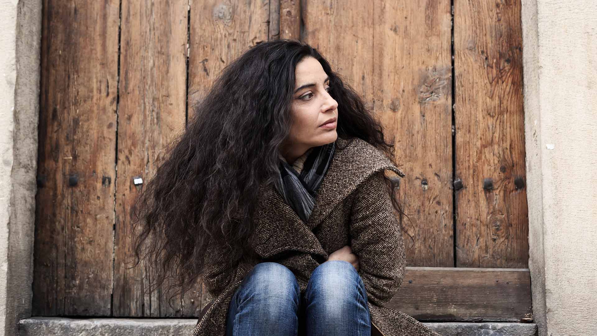 Beautiful young woman with long hair and coat sitting on the stairs at the door, thinking and looking around.