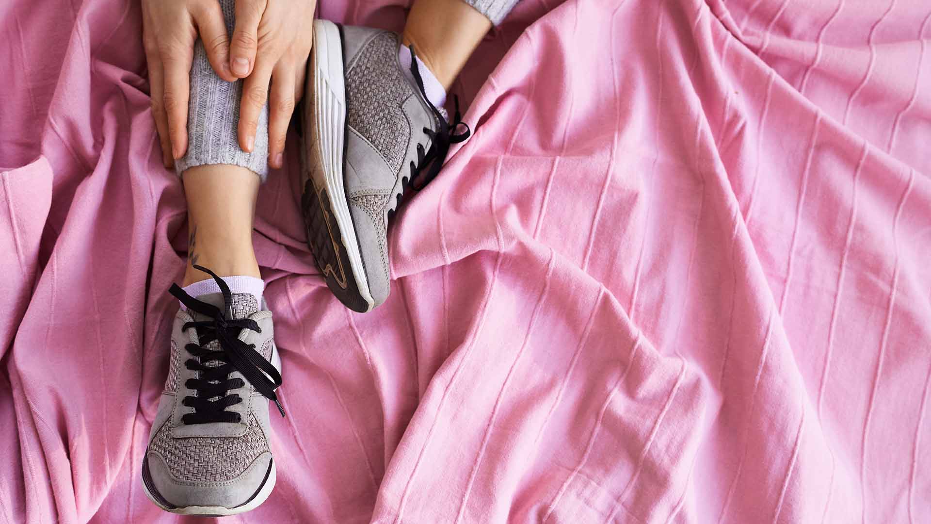 High angle view of female athlete in wool sport pants and sneakers sitting on bed, touching her ankle with both hands and stretching