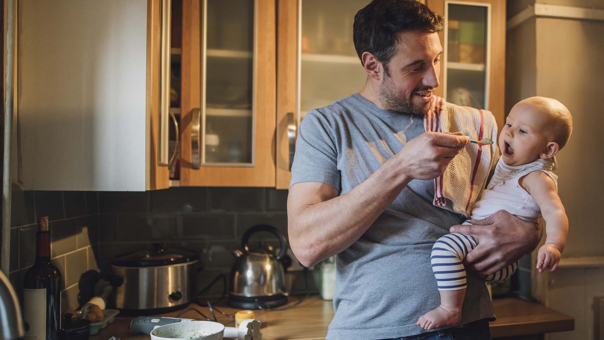 Father is standing in the kitchen of his home with his baby in his arms. He is feeding him with a spoon and spinach and vegetables can be seen on the worktop with a blender.