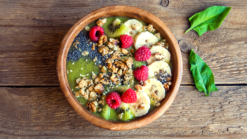 Healthy breakfast green smoothie bowl topped with fruits, nuts, berries and seeds over rustic wooden background