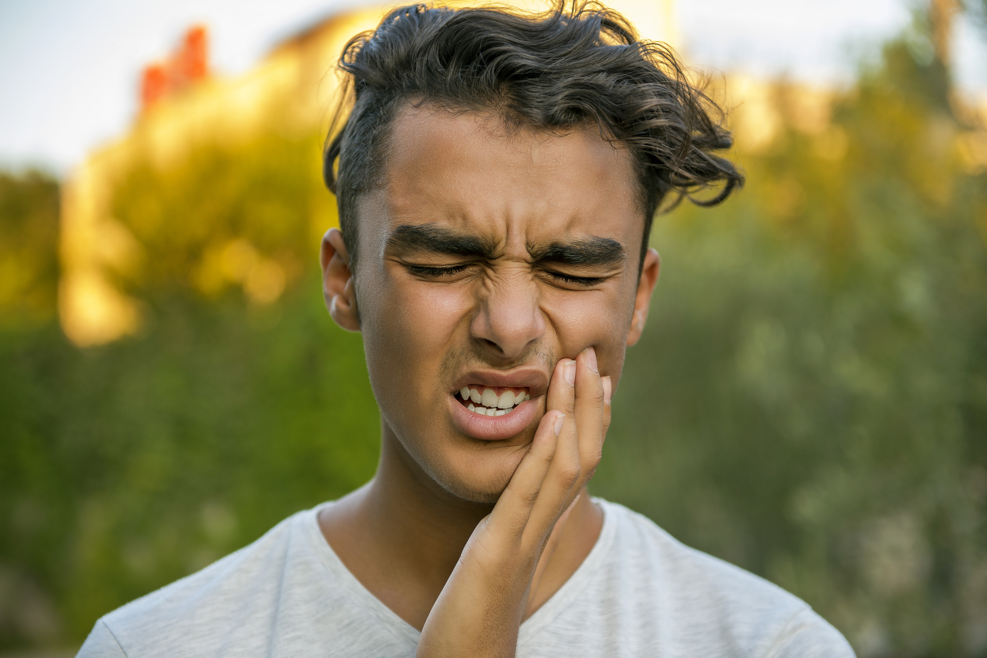 Worried teenage boy rubbing his mouth because of toothache. Portrait of a young man standing in front of tree area and closing his eyes in pain because of toothache. There are trees behind man. Horizontal composition. Image taken with Nikon D800 and developed from RAW format.