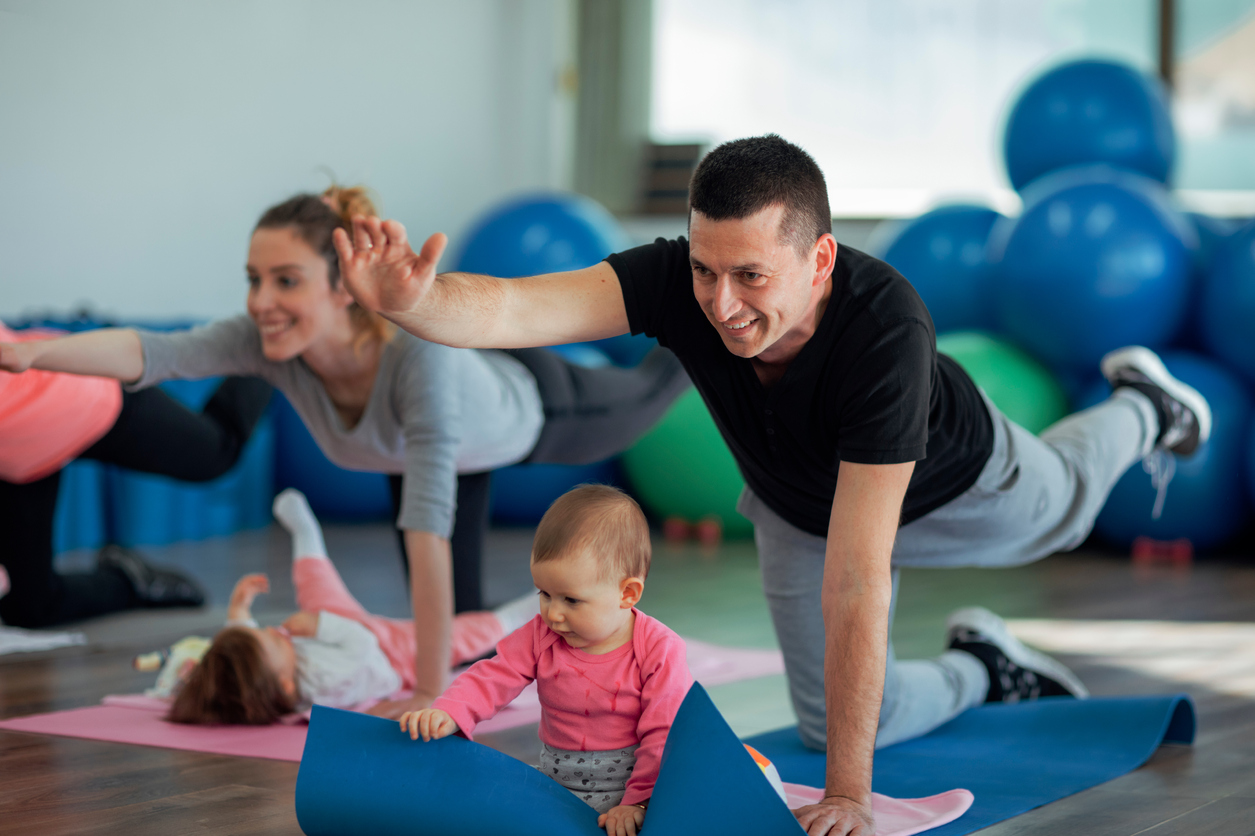Parents Exercising with Their Babies in a Gym. Selective focus on father exercising with his baby. He is exercising on floor on exercising mat and baby is sitting in front of her dad. He is happy to exercise together with his baby and stay fit.