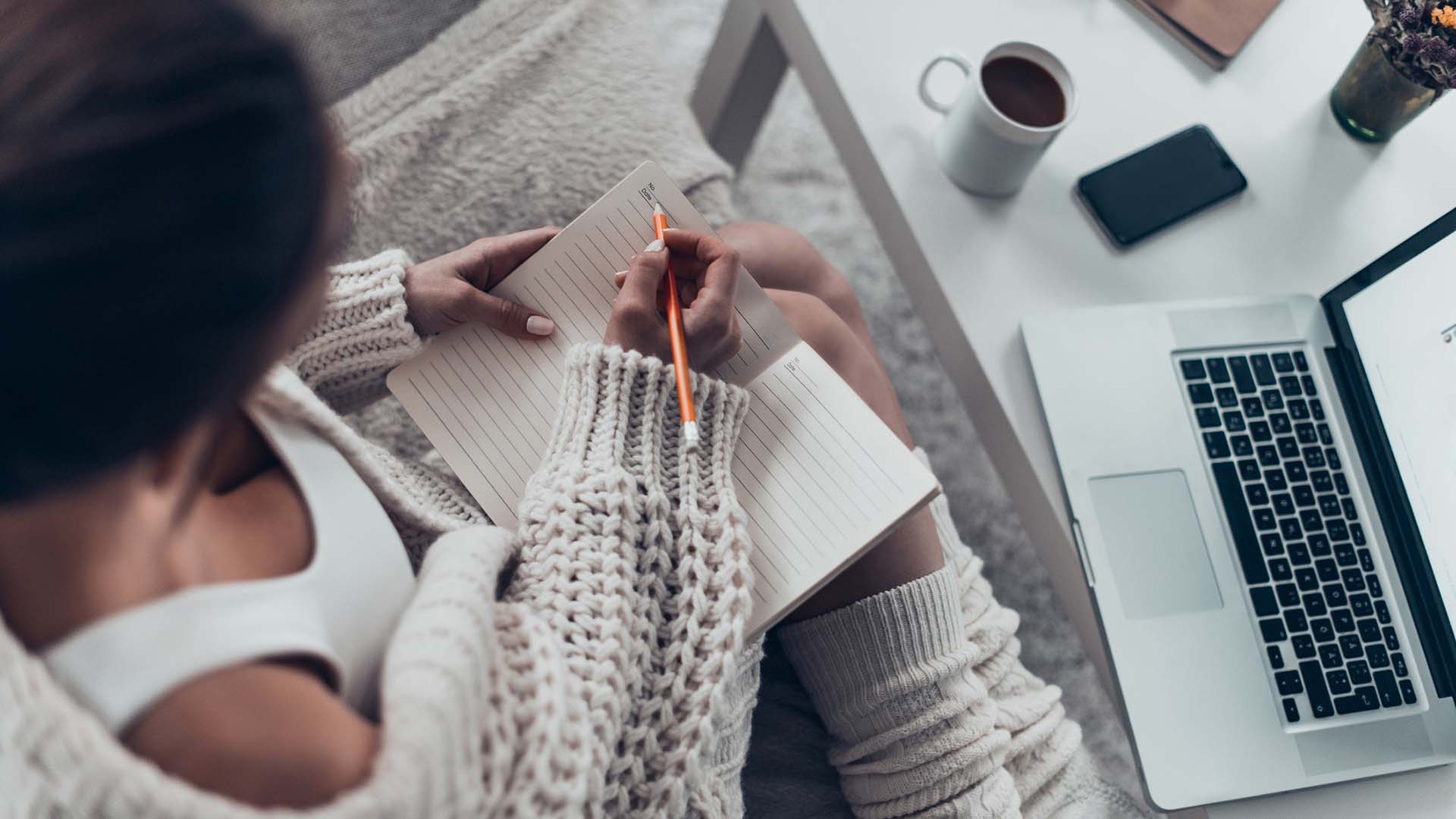 Top view of attractive young woman writing something in her note pad while spending weekend at home