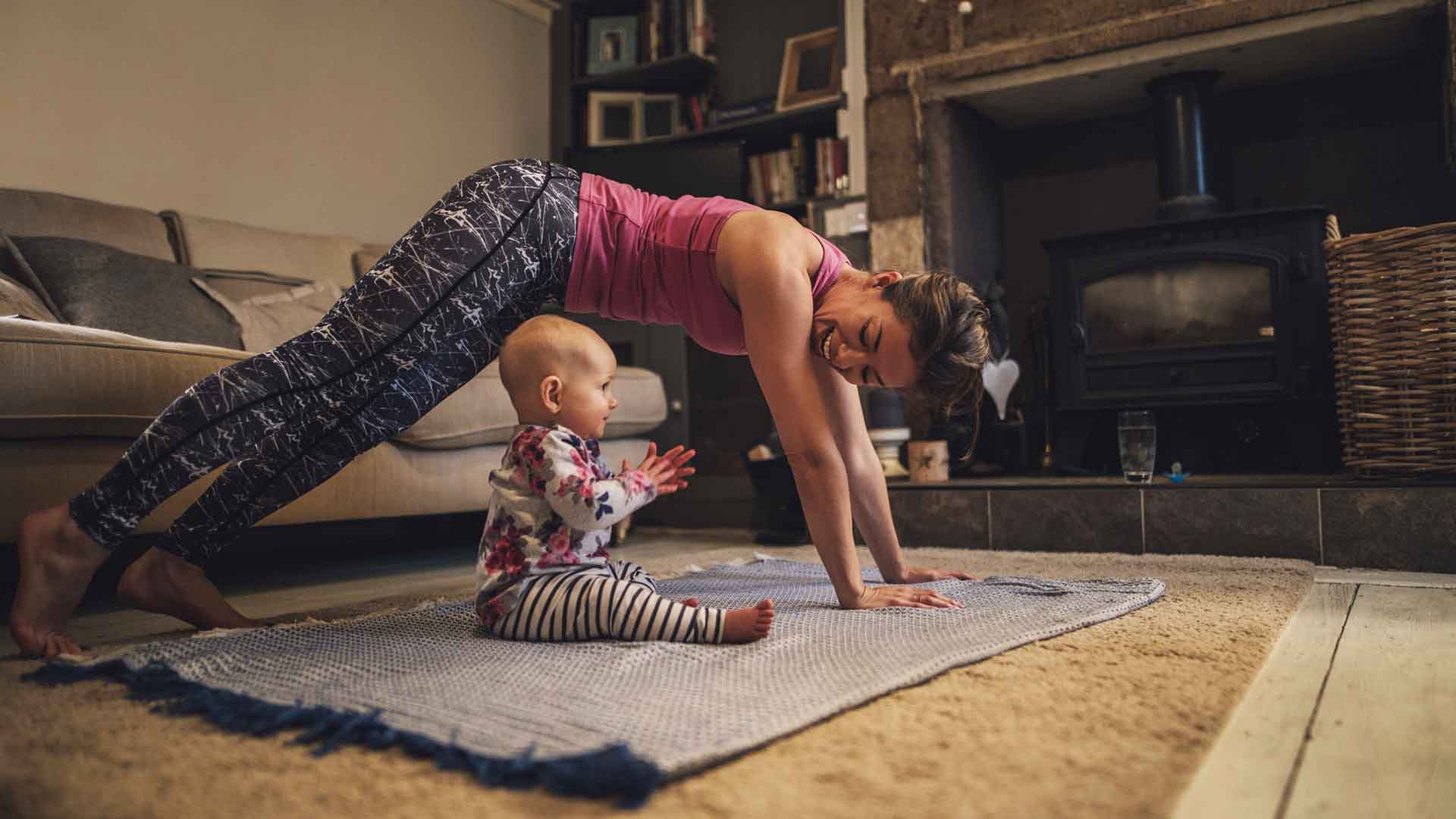 Young mother is practicing yoga in the living room of her home. Her baby daughter is sitting next to her trying to copy her.
